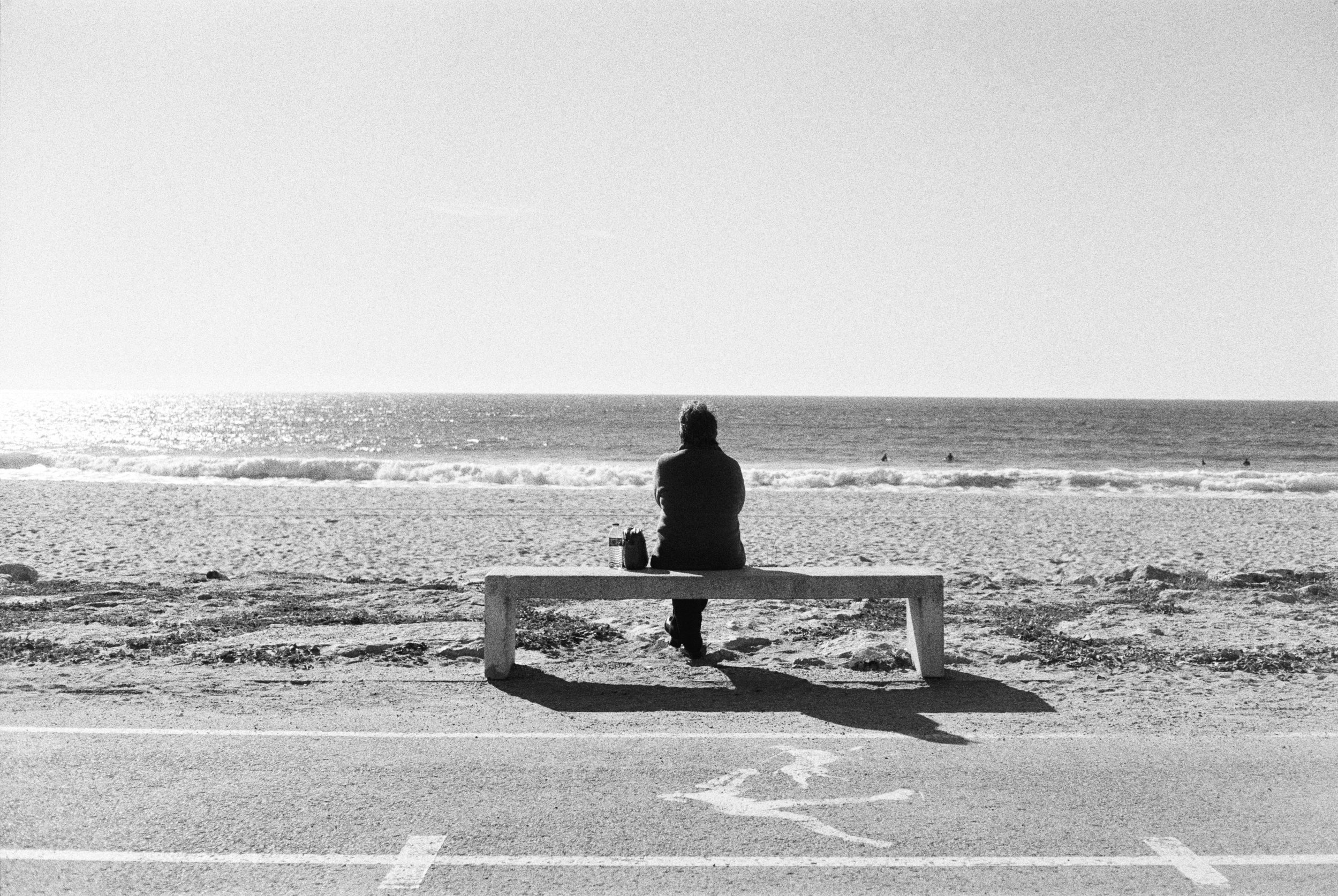 A person sitting on a bench facing the ocean at the beach, with surf and a few surfers in the water, black and white photo.