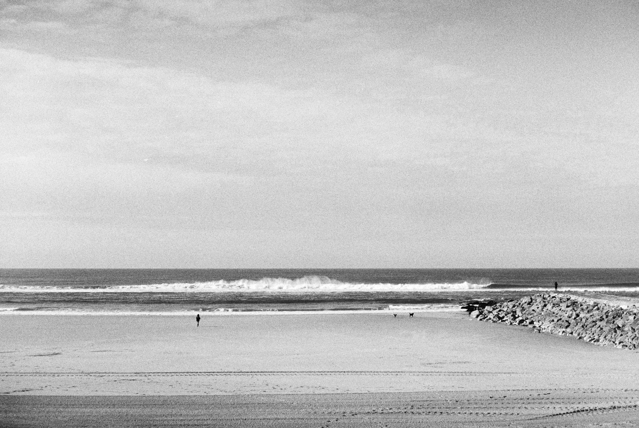 Black and white photo of a beach with a rocky jetty on the right. Two people are standing on the sand, and the ocean waves are crashing in the background under a mostly clear sky.