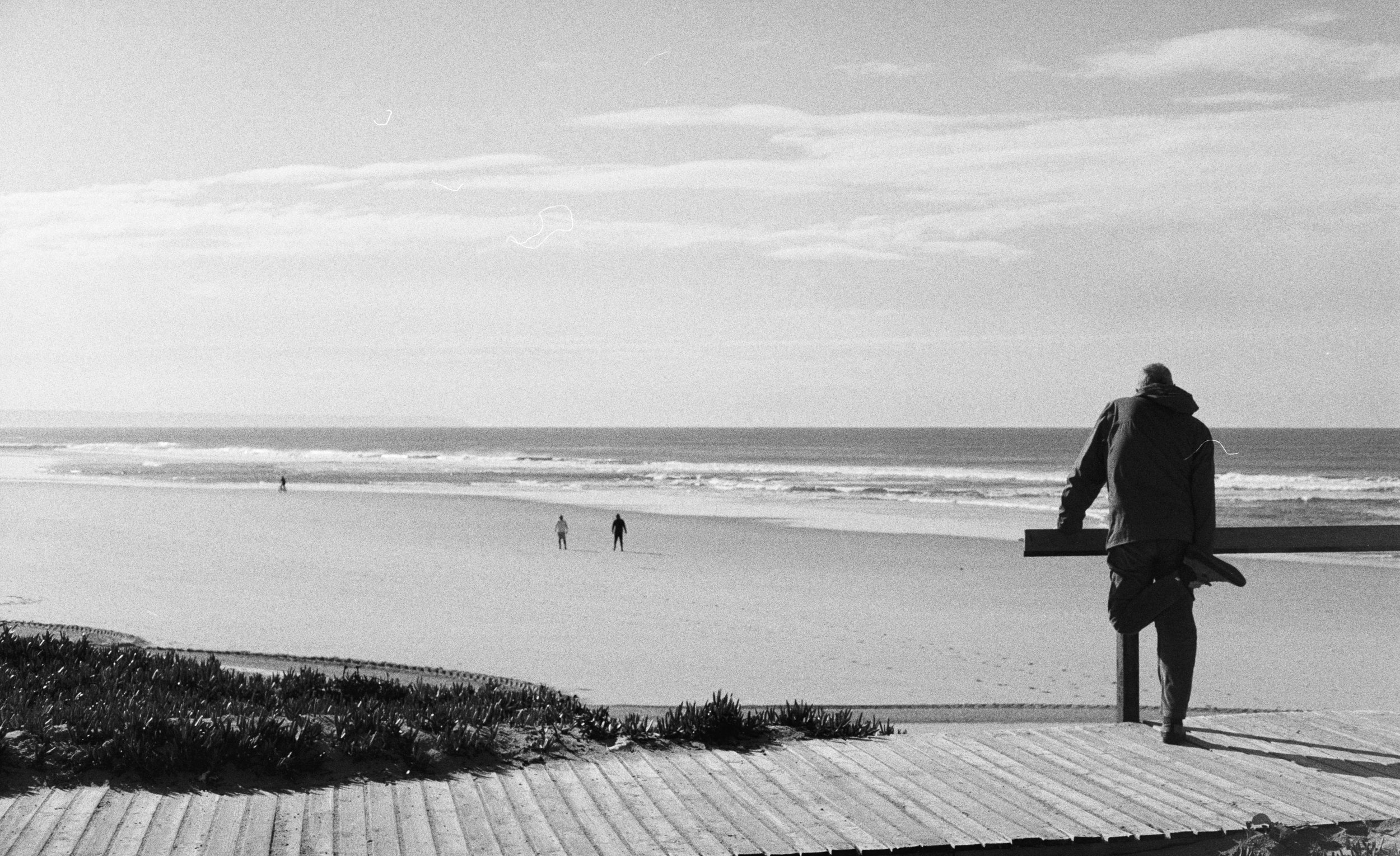 A black-and-white photo of a person in a hoodie holding a surfboard, standing on a wooden walkway and looking at the ocean, with three other people walking in the distance on the beach.