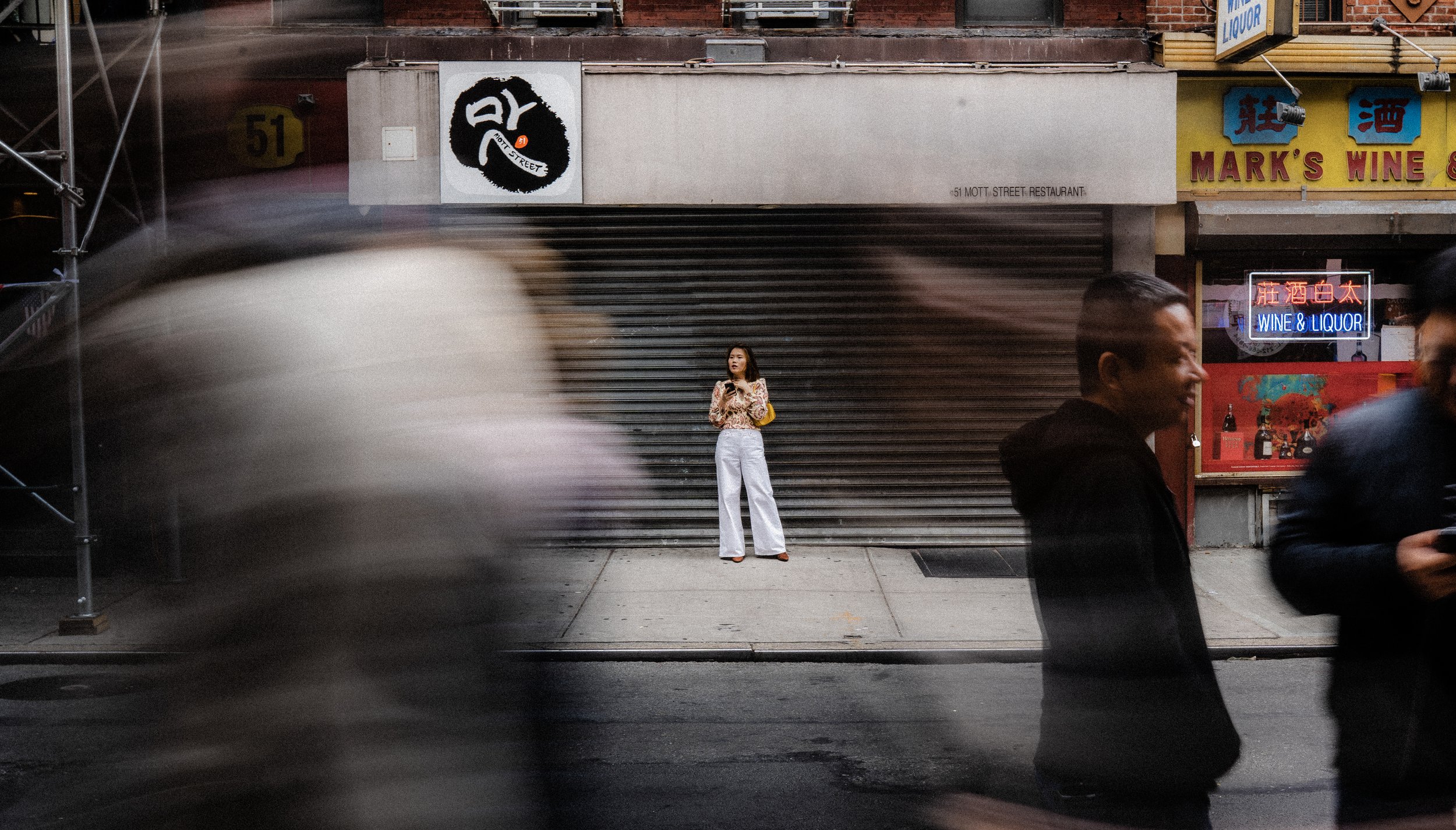 A young woman stands alone on a city sidewalk in front of a closed storefront with metal security shutters, while blurred pedestrians walk past in the foreground and background, with neon signs and storefronts visible.