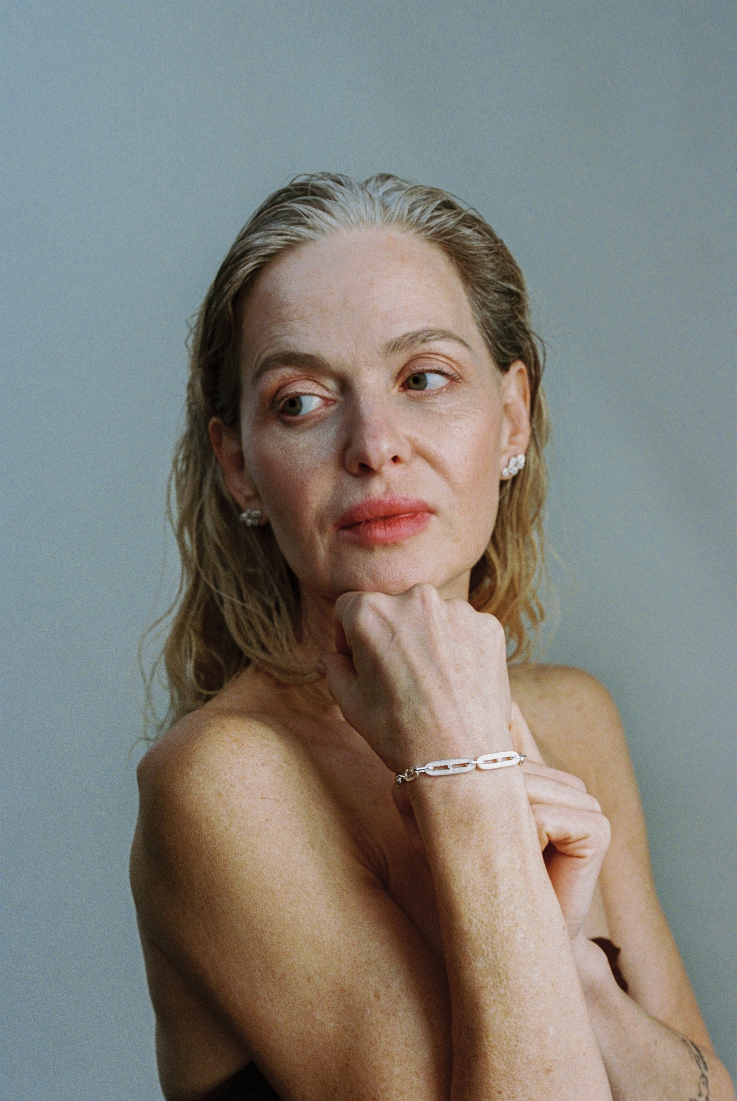 A woman with shoulder-length blonde hair, light skin, and minimal makeup, resting her chin on her hand while looking to the side, against a plain, muted background.