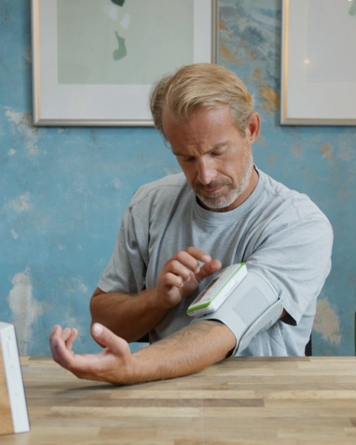 A man in a gray t-shirt is sitting at a wooden table, taking his blood pressure with a digital monitor, in a room with blue painted walls and framed artwork.
