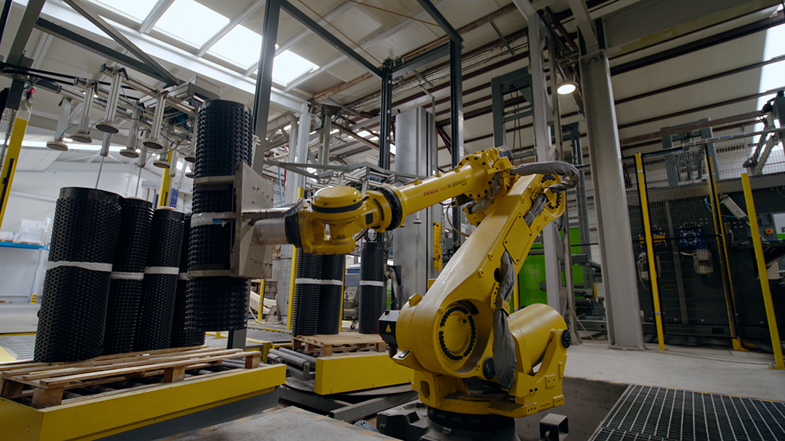 A yellow industrial robotic arm in a manufacturing facility handling black textured cylindrical objects on wooden pallets.