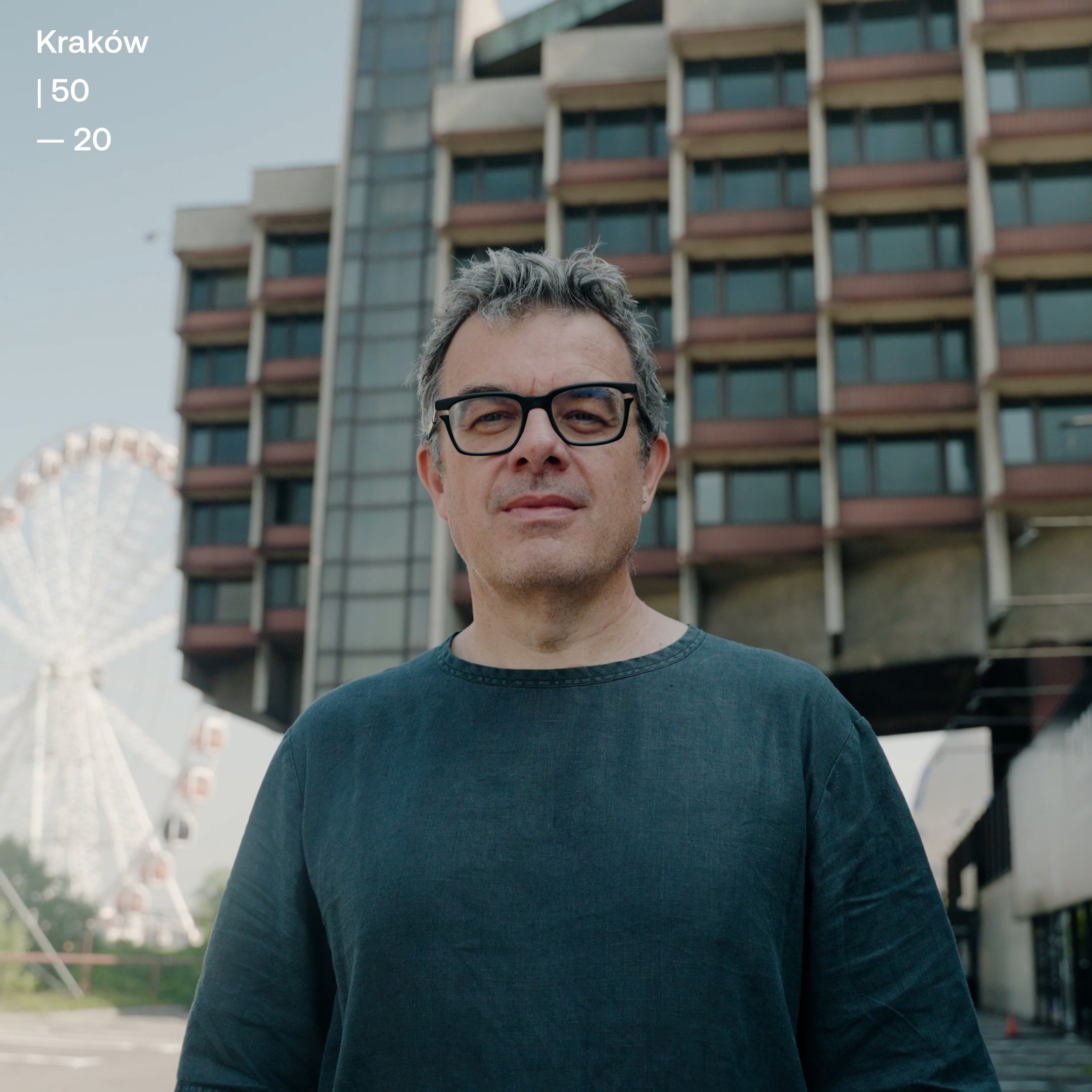 A middle-aged man with gray hair and black glasses stands outdoors in front of a building with a grid-like window pattern. In the background, there is a Ferris wheel and an amusement park.