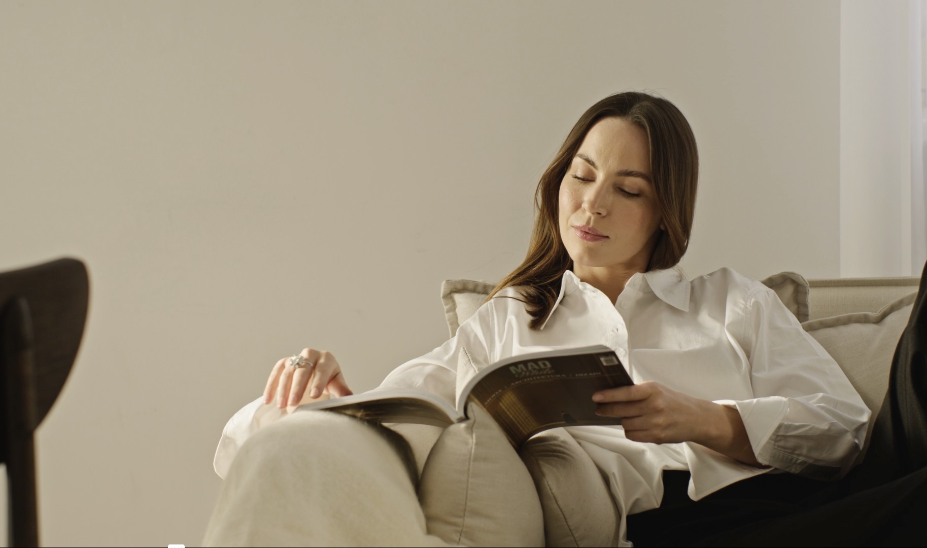A woman with brown hair wearing a white blouse sitting on a beige couch, reading a magazine indoors.
