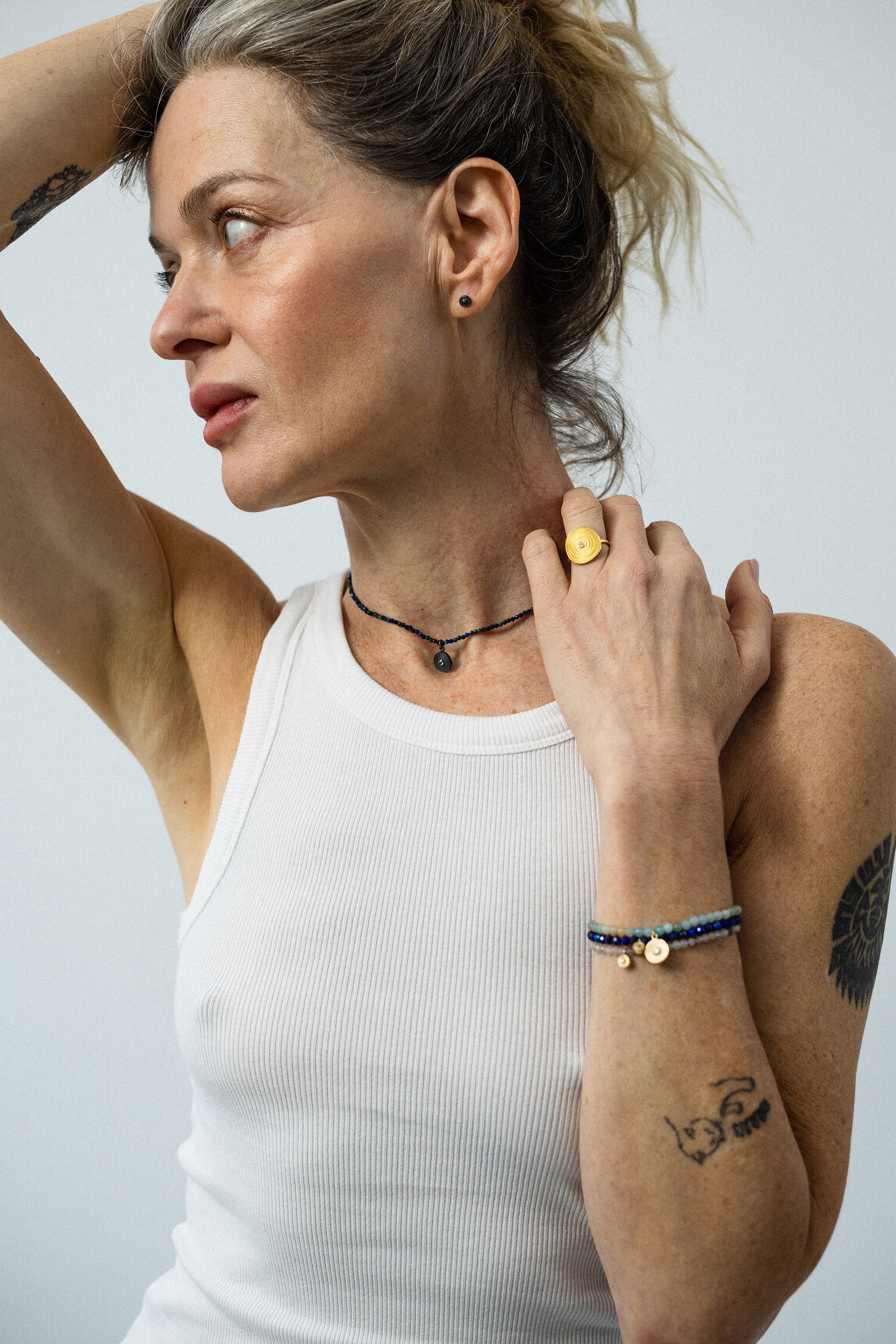 A woman with tattoos on her arm, wearing a white tank top, black necklace, yellow ring, and beaded bracelet, has her left arm raised behind her head and touches her neck.