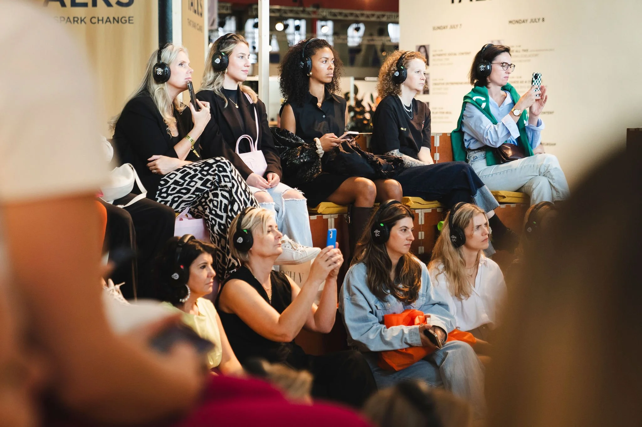 Group of women seated and listening at an event, all wearing headphones, with some taking photos or videos with their phones.
