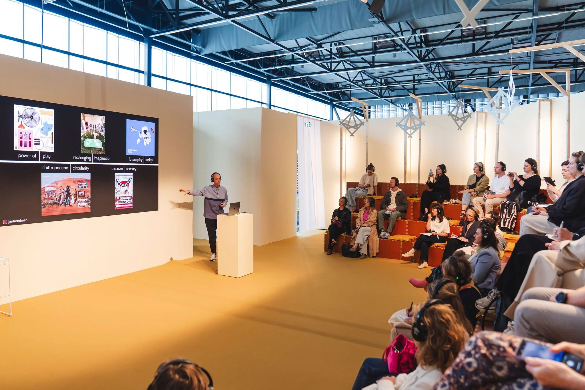 A presenter stands at a podium in front of a large screen showing colorful thumbnails and text, addressing an audience seated on tiered orange seating in a brightly lit, modern conference room with high ceilings and large windows.