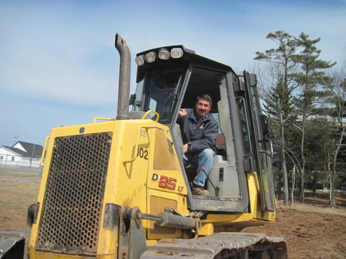 A man, a volunteer with Acadia Park and Recreation Centre sitting inside a yellow bulldozer on a construction site with a few trees and residential buildings in the background.