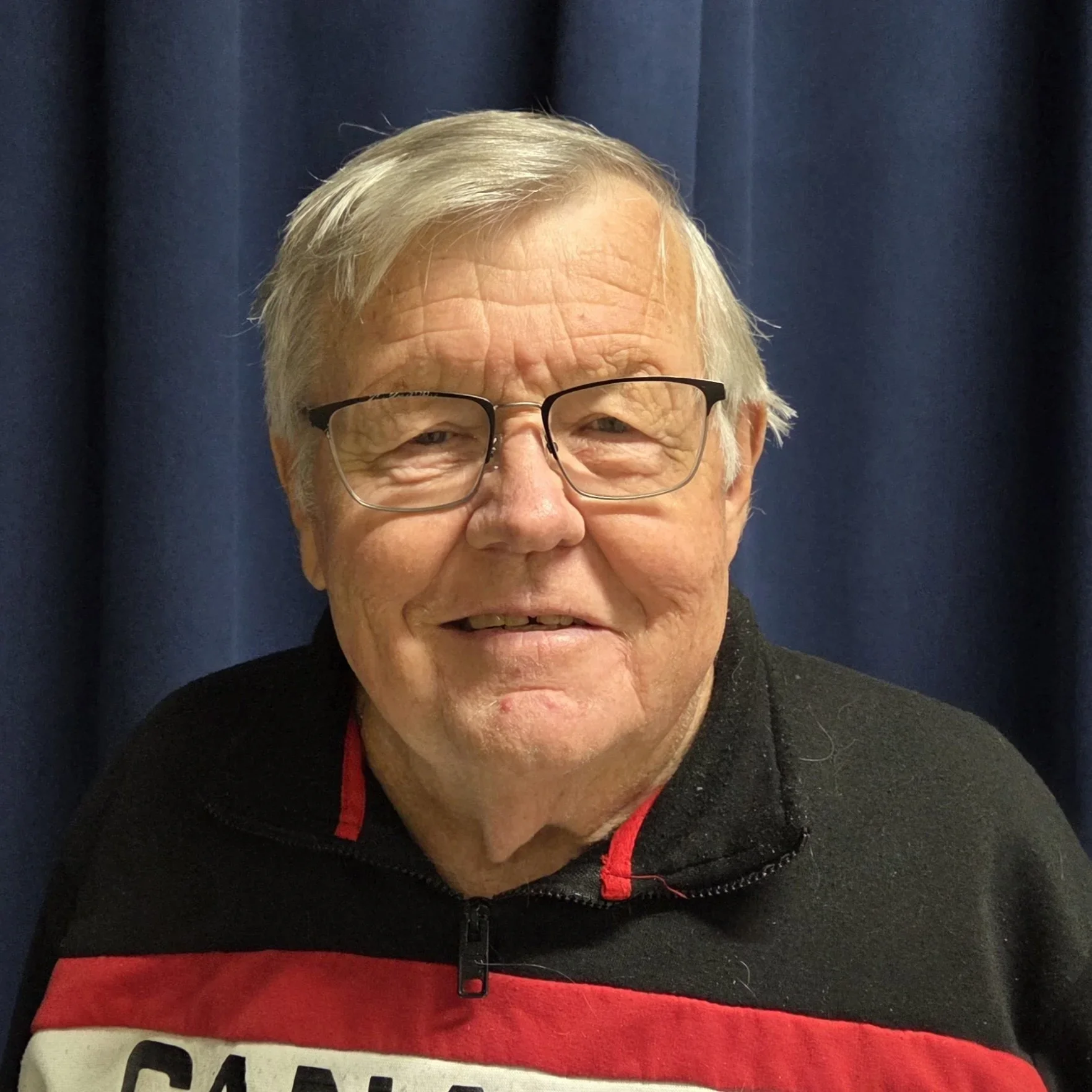 A man with glasses and gray hair smiling, wearing a black and red jacket, standing in front of a dark blue curtain.
