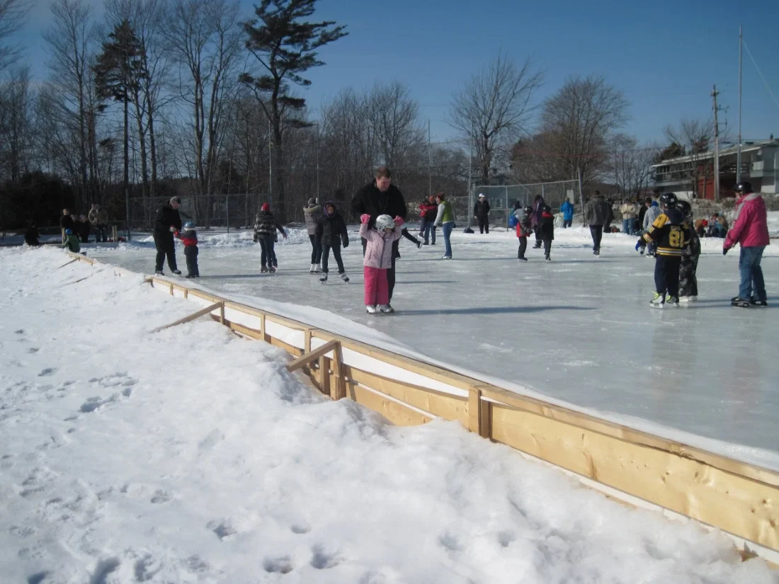 People, including children, skating on an outdoor ice rink in winter at Acadia Park in Sackville, NS, surrounded by snow and leafless trees under a blue sky.