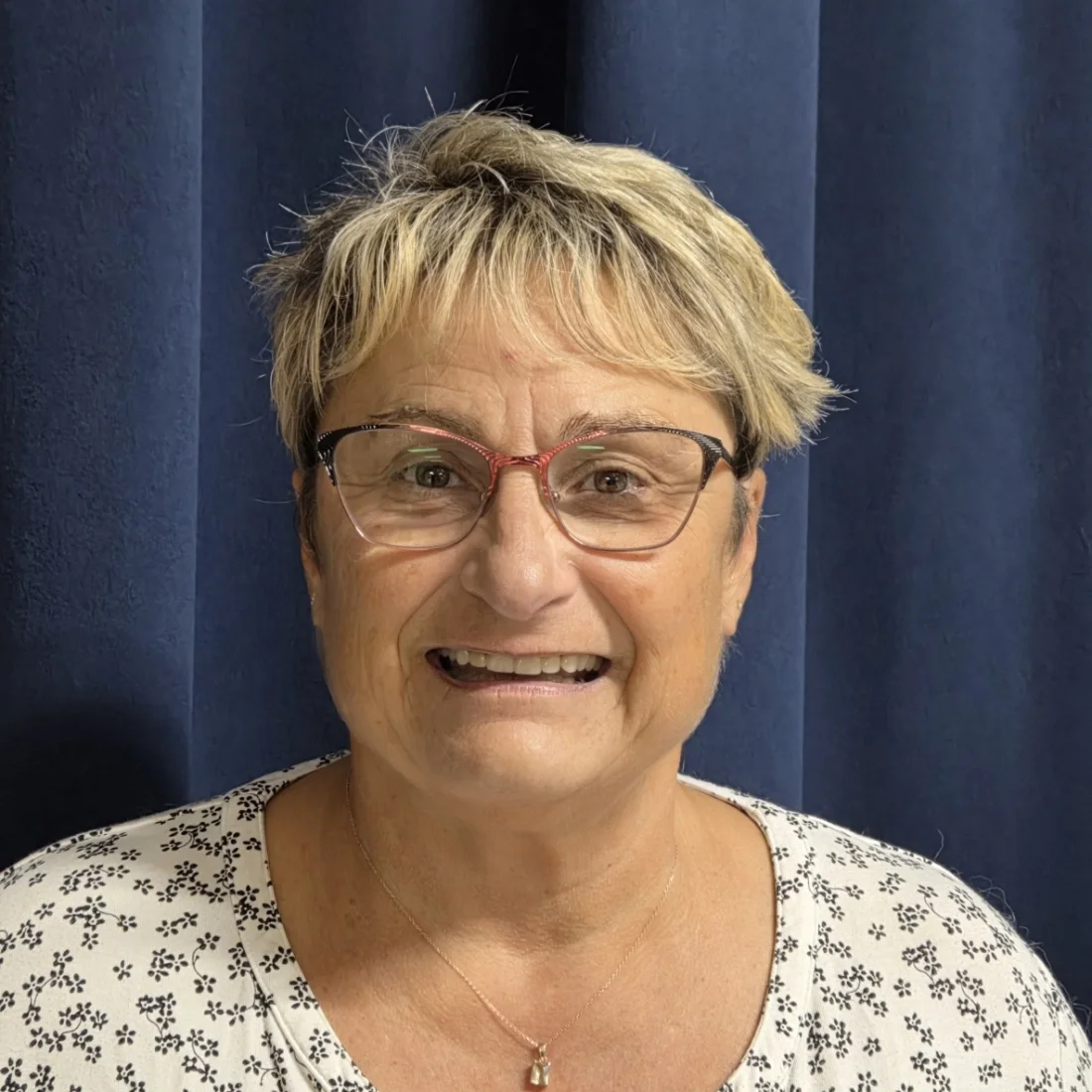 A smiling woman with short blonde hair, wearing glasses, a floral patterned top, and a necklace, standing in front of dark blue curtains.
