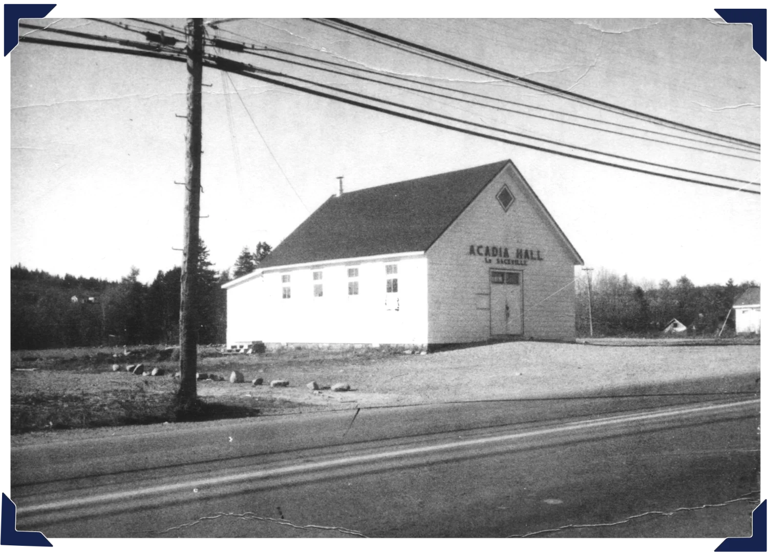 Acadia Hall in the 1940s, located near a road with overhead power lines, trees in the background, and a gravel area in front.