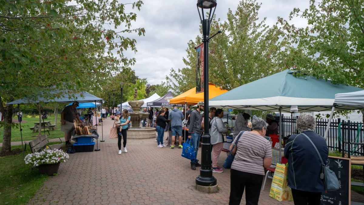 People browsing vendors at the outdoor Sackville, NS Farmer's Market with tents, trees, and a fountain in the background.