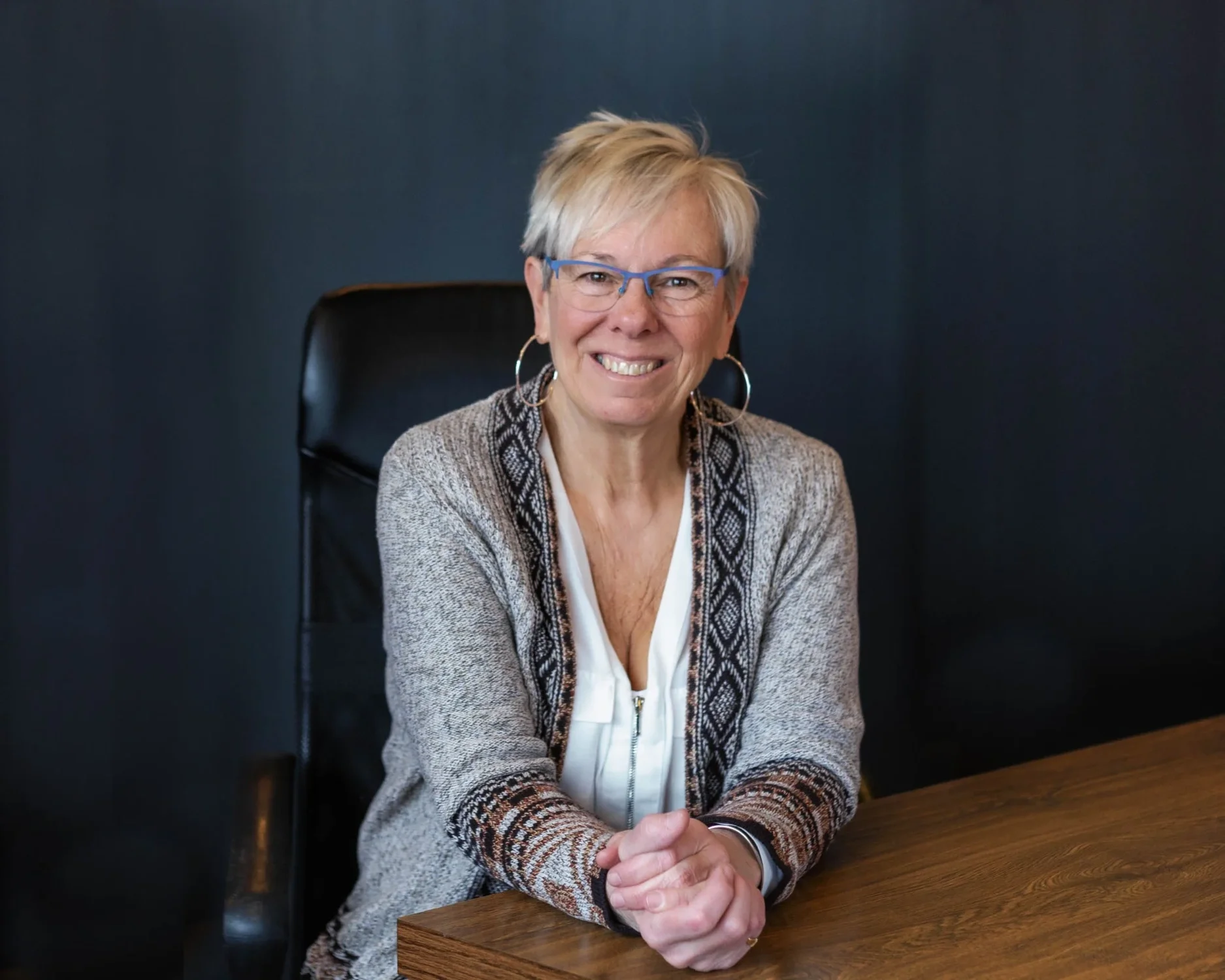 A smiling woman with short gray hair, glasses, and hoop earrings sitting at a wooden desk against a dark background.