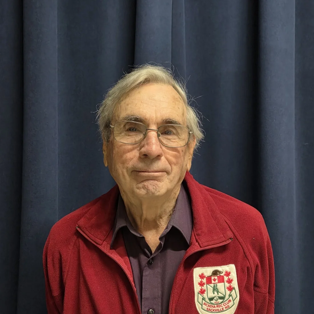 A man with glasses wearing a maroon jacket with a crest that reads 'ACADIA RECREATION CLUB SACKVILLE NS' standing in front of a blue curtain.
