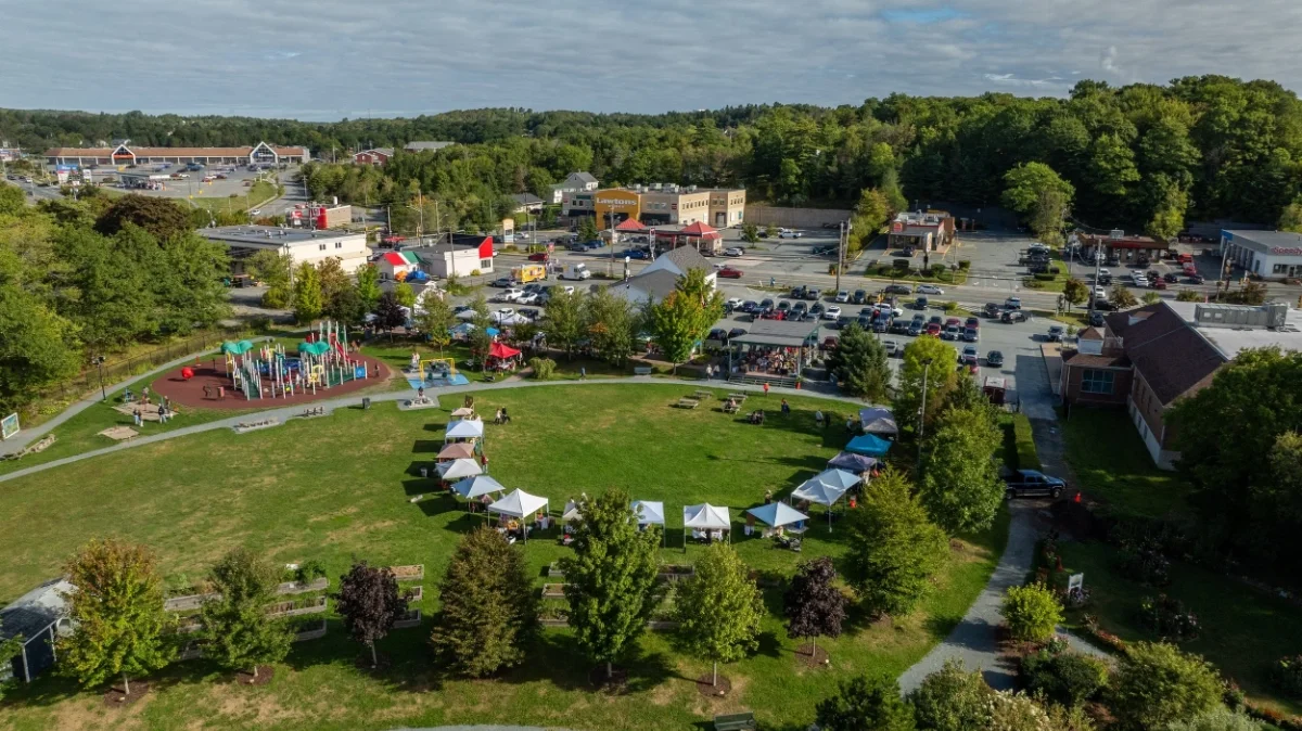 Acadia Park in Sackville, NS with a playground, tents, and picnic tables, surrounded by trees and near a shopping center with cars parked and stores in the background.