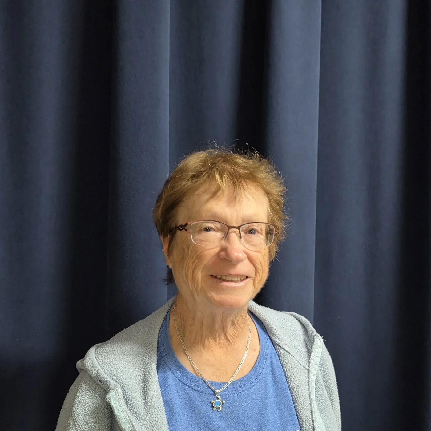 Smiling woman with glasses and short hair standing in front of dark blue curtain.