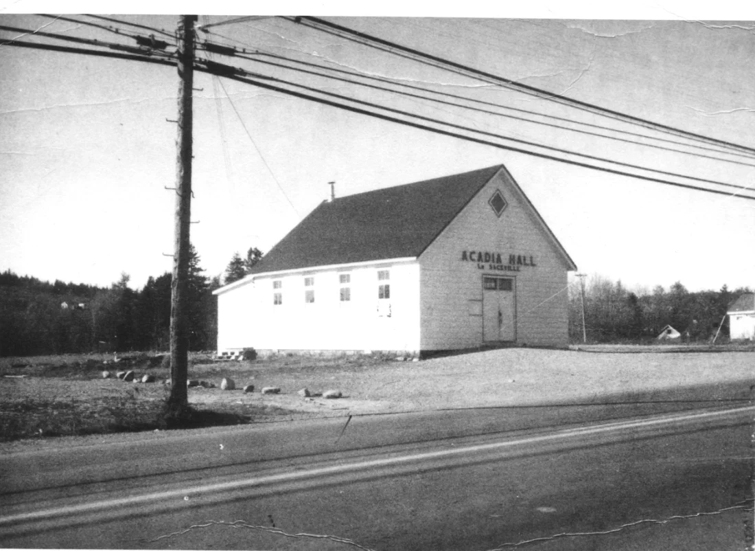 Acadia Hall in the 1940s. There is a road in the foreground and power lines overhead. The hall has a gabled roof and small windows.