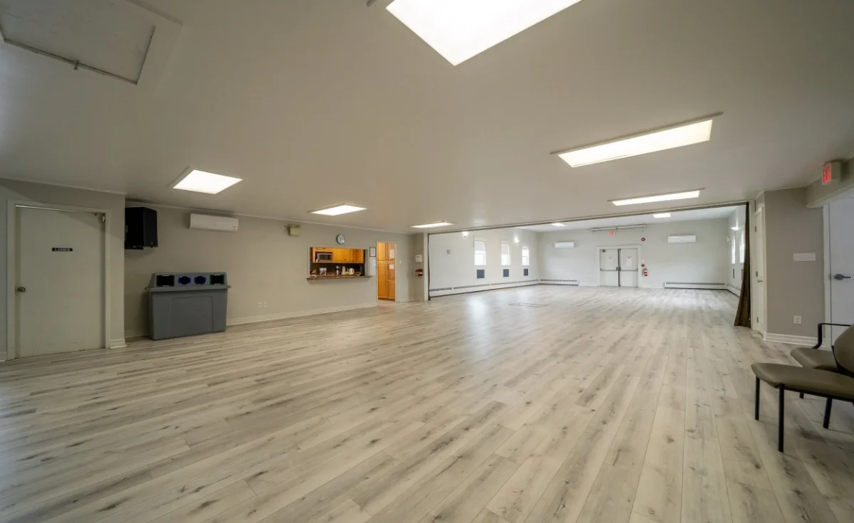 Empty large room in Acadia Hall in Sackville, NS with light wood flooring, white walls, and ceiling; several windows, closed doors, and minimal furniture, including chairs and a counter.
