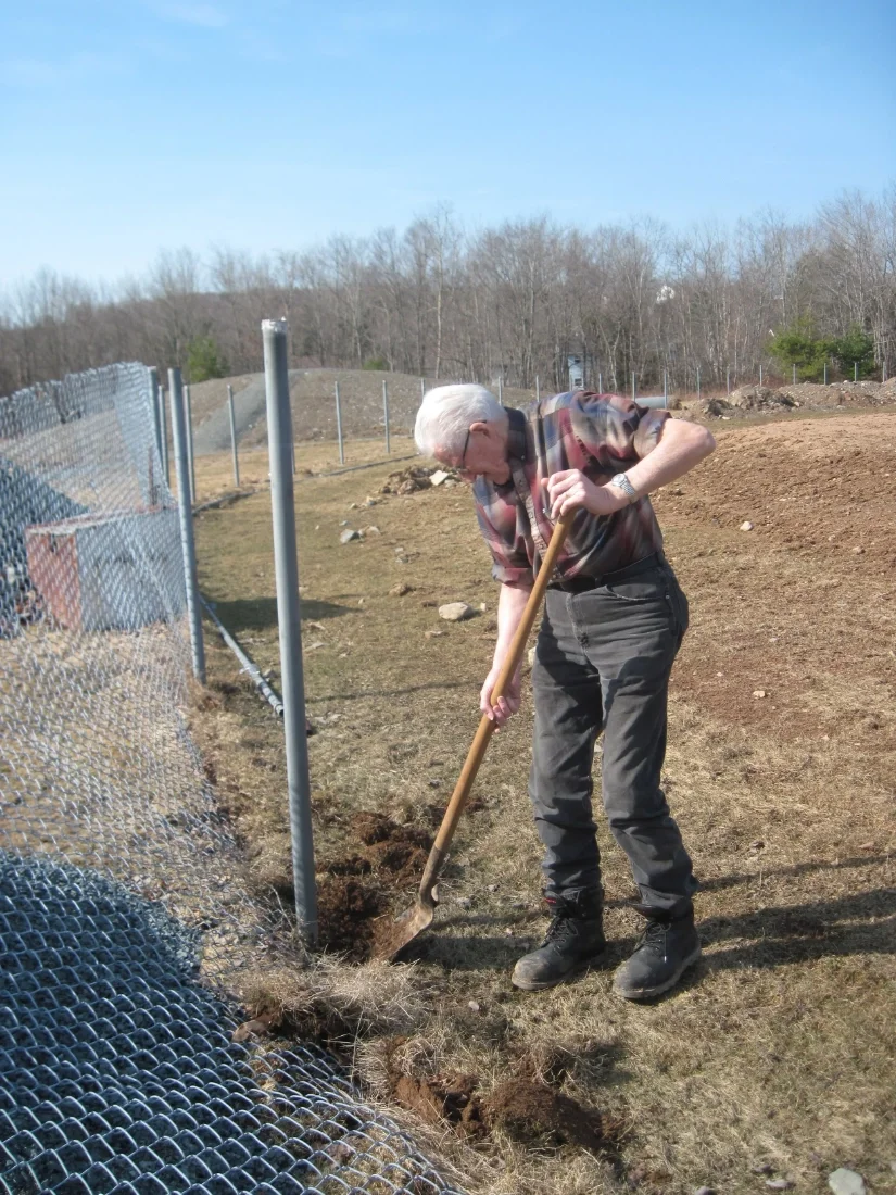 A man with white hair wearing glasses, a patterned shirt, dark pants, and boots, is digging up one of Acadia Park's baseball field's chain-link fence with a shovel outdoors on a sunny day. There are trees and a grassy field in the background.
