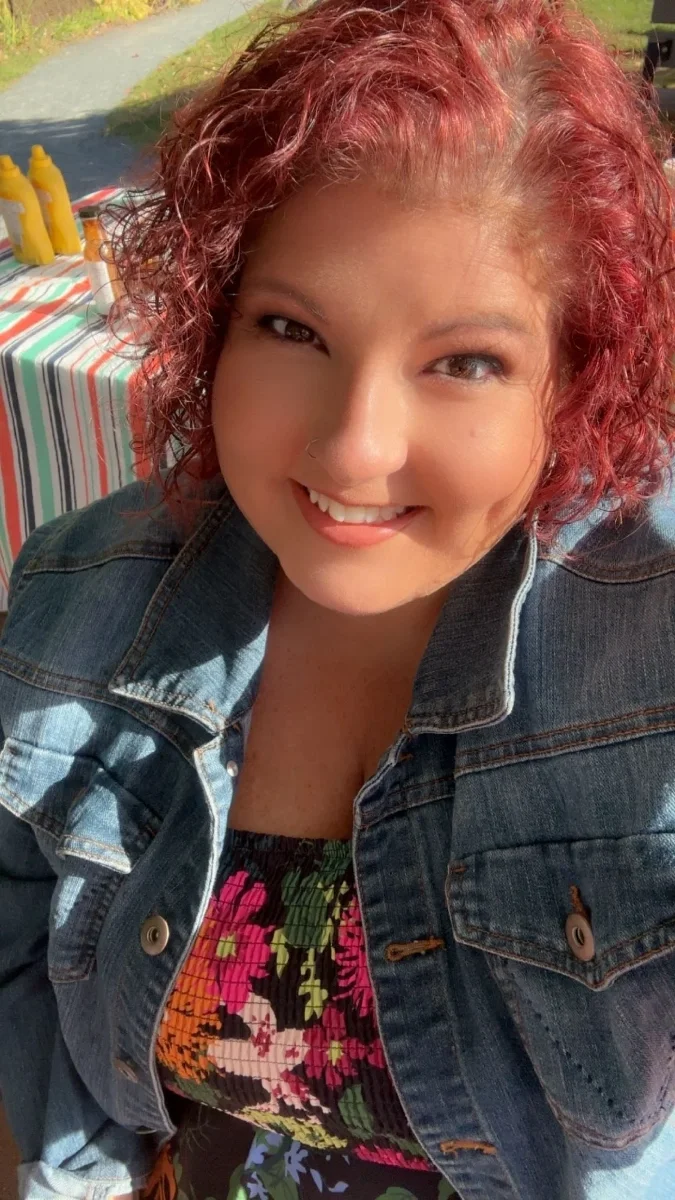 A woman with curly red hair smiling at the camera outdoors during daytime, wearing a denim jacket and a colorful floral top.