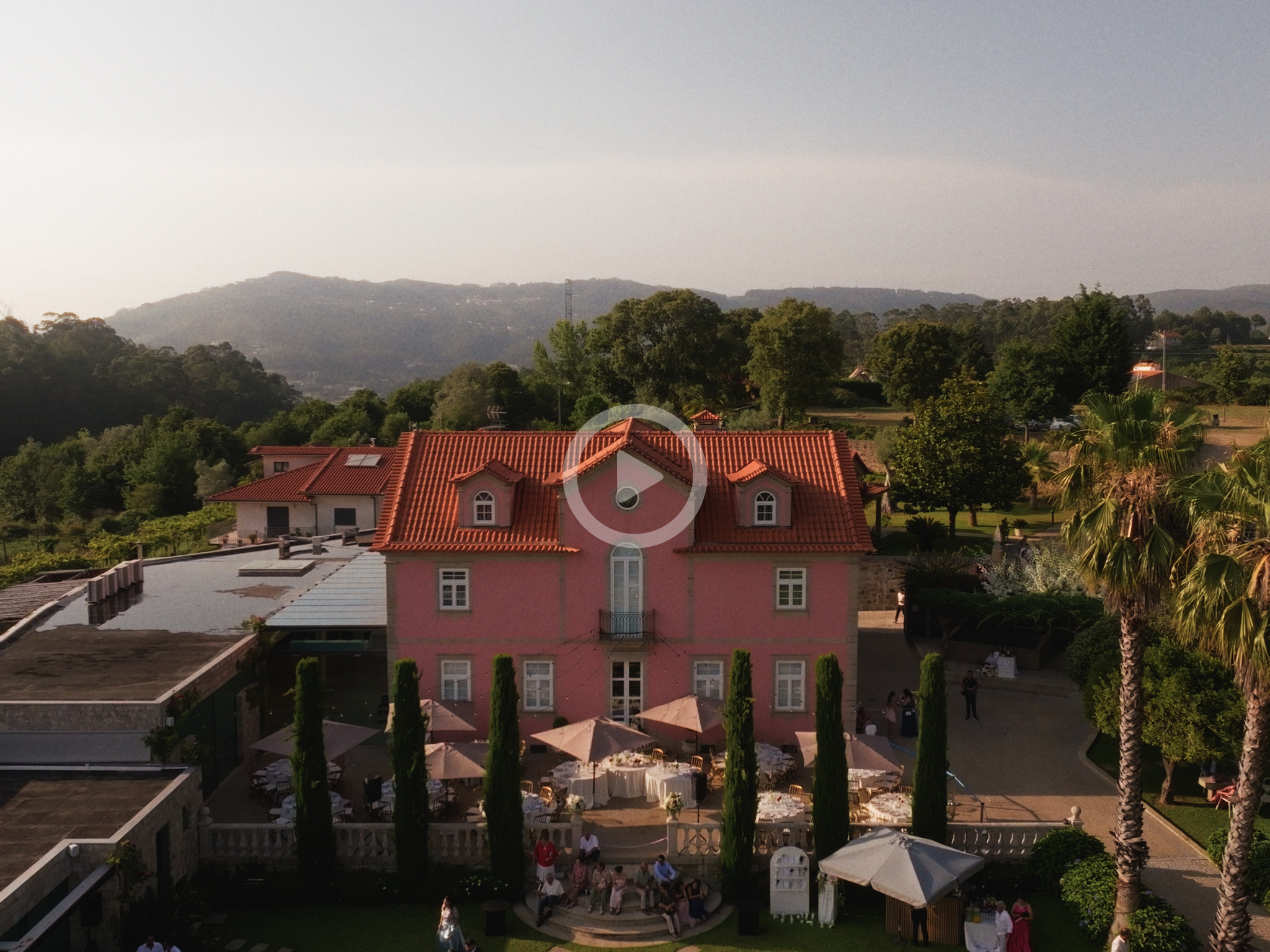 A landscape view of a residential area with trees, a pink house with a red roof, and an outdoor event setup with tables, umbrellas, and people dressed in formal attire, possibly for a wedding or celebration, with mountains in the background and a clear sky.