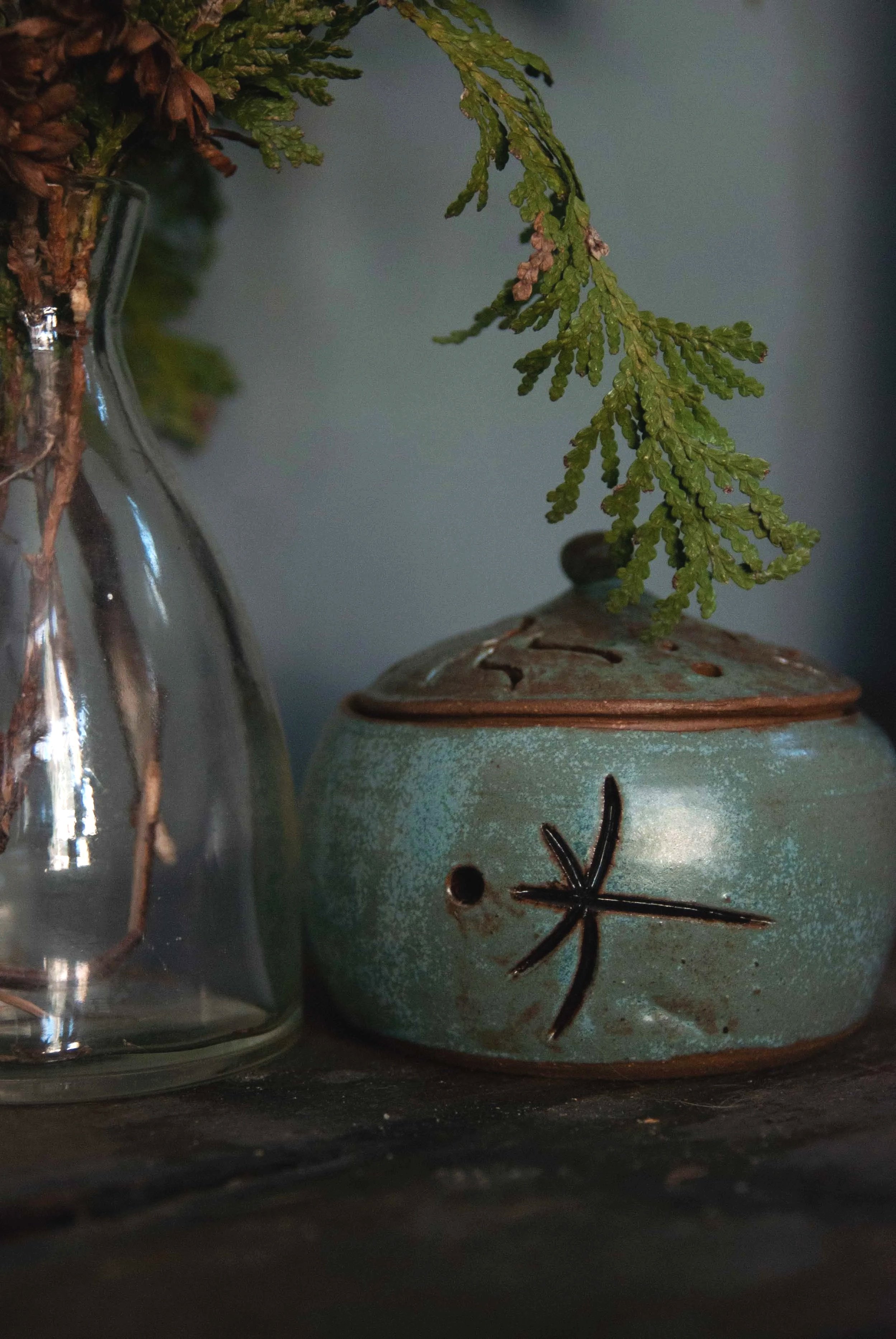 Close-up of a ceramic incense burner with a custom sigil/symbol design, next to a glass vase with green foliage against a dark background.