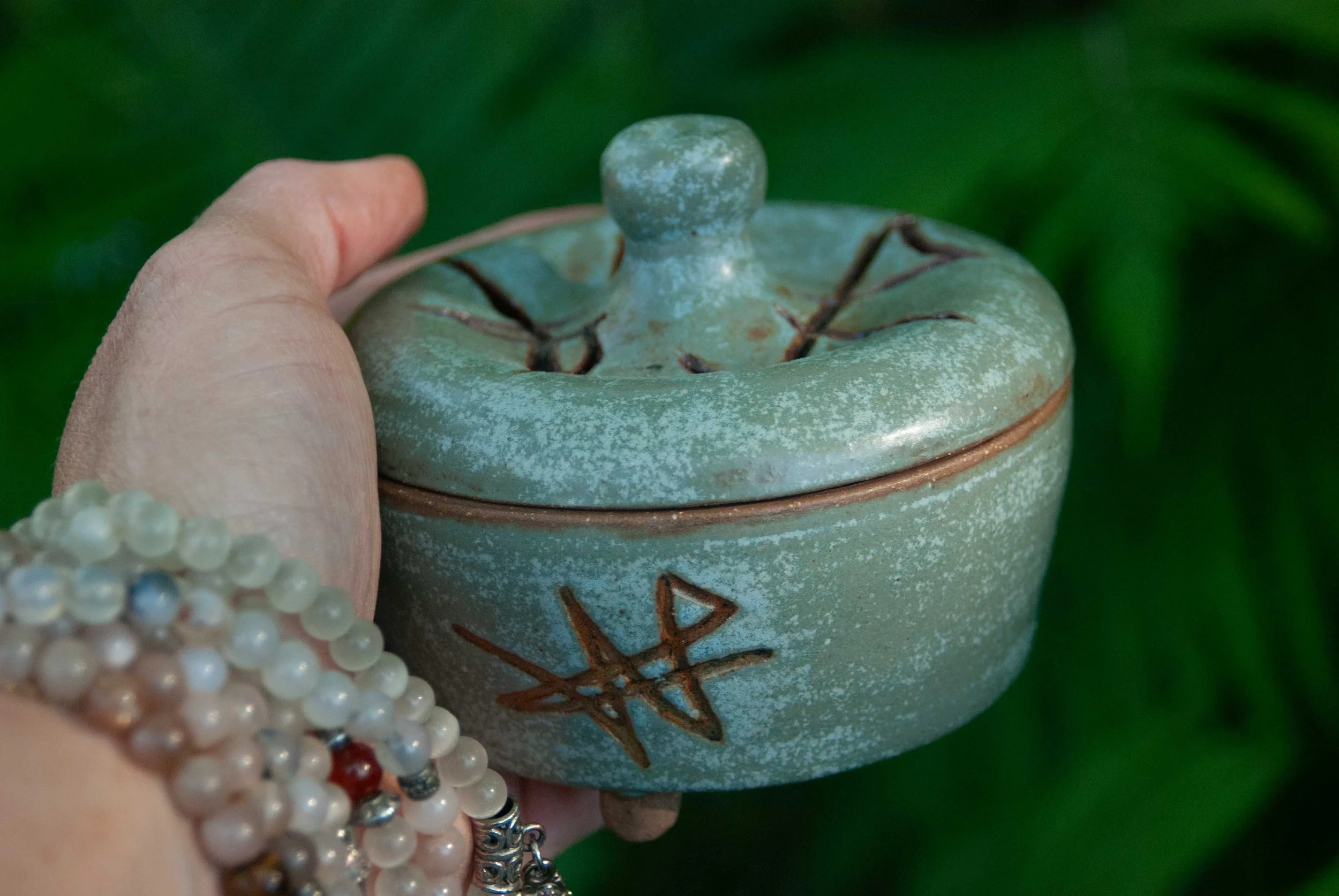 Close-up of a green ceramic container with a lid, decorated with a copper-colored sigil, a custom symbol. A person's hand and a mala bracelet are partially visible in the foreground, with a blurred green background.