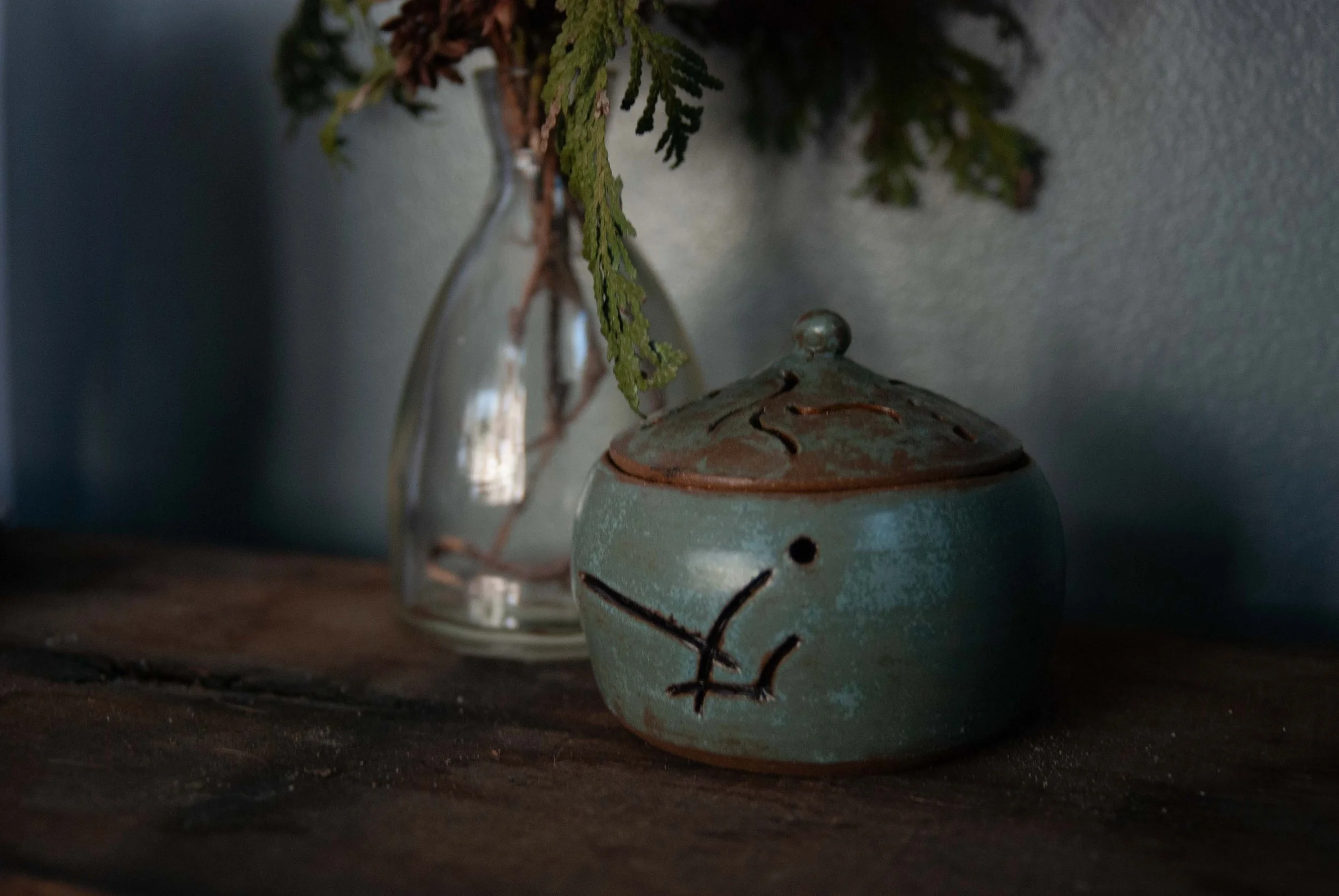 A small ceramic incense burner with a lid and a sigil carving, placed on a wooden surface with a glass vase and green foliage in the background.