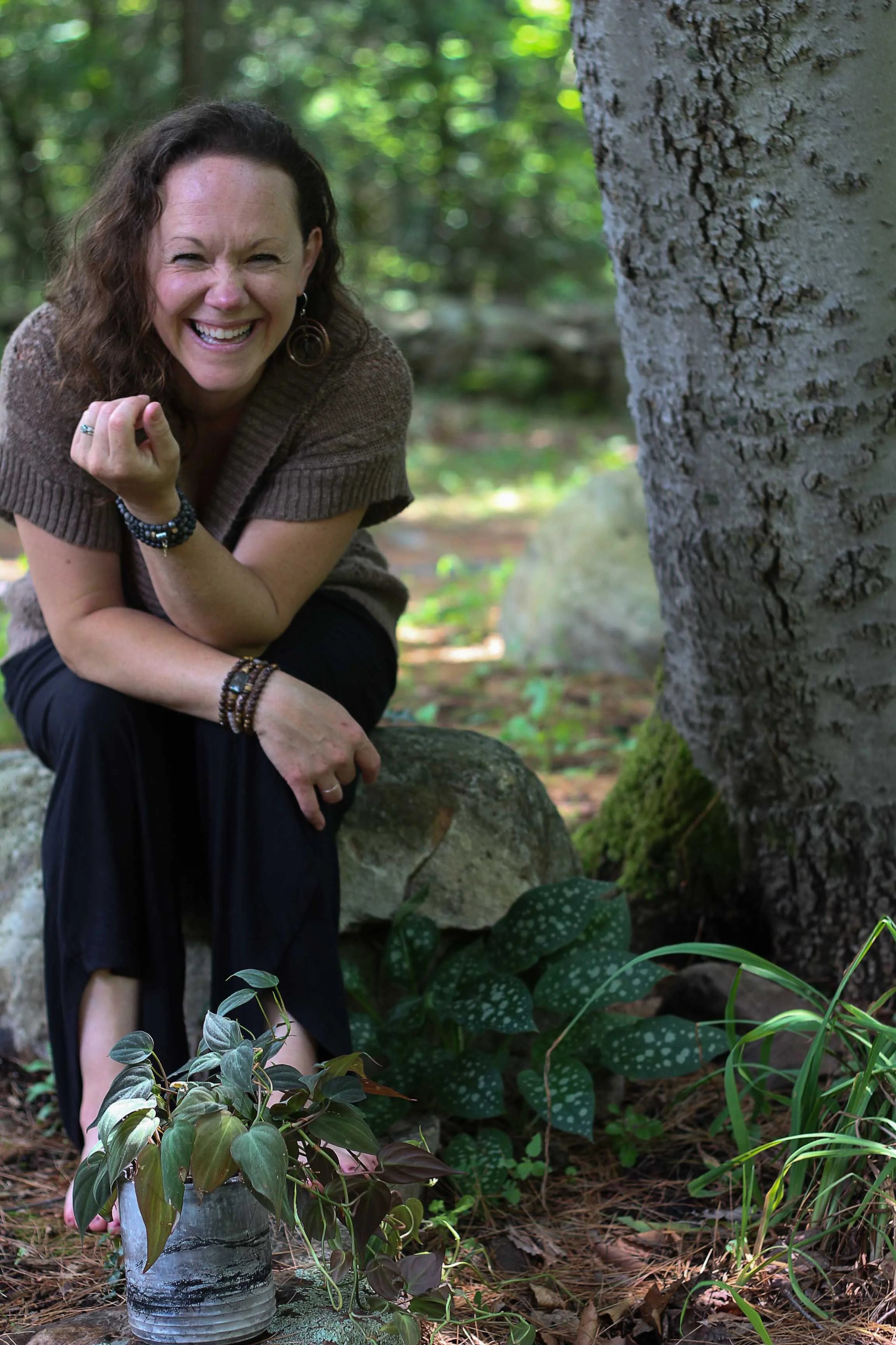 Artist of Potted Earth crouching near a tree in a forest, smiling and laughing, surrounded by green plants and rocks.