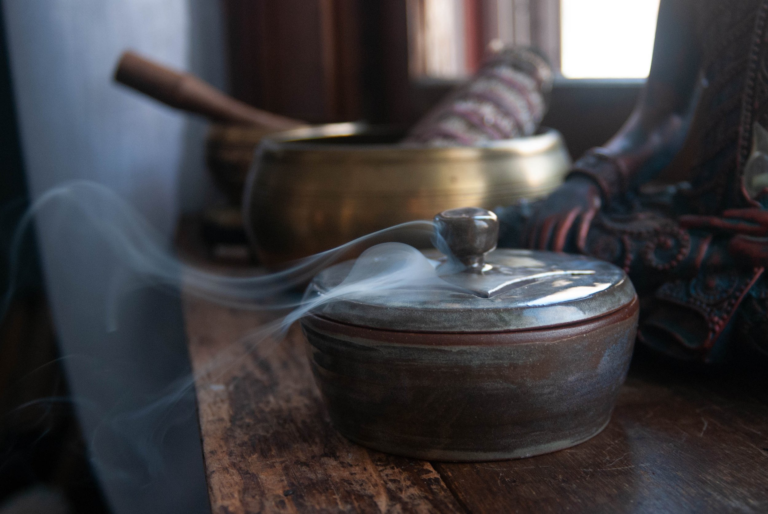 An artisan ceramic bowl with a lid and a carved sigil (symbol), emitting incense, placed on a wooden surface in a cozy rustic setting.