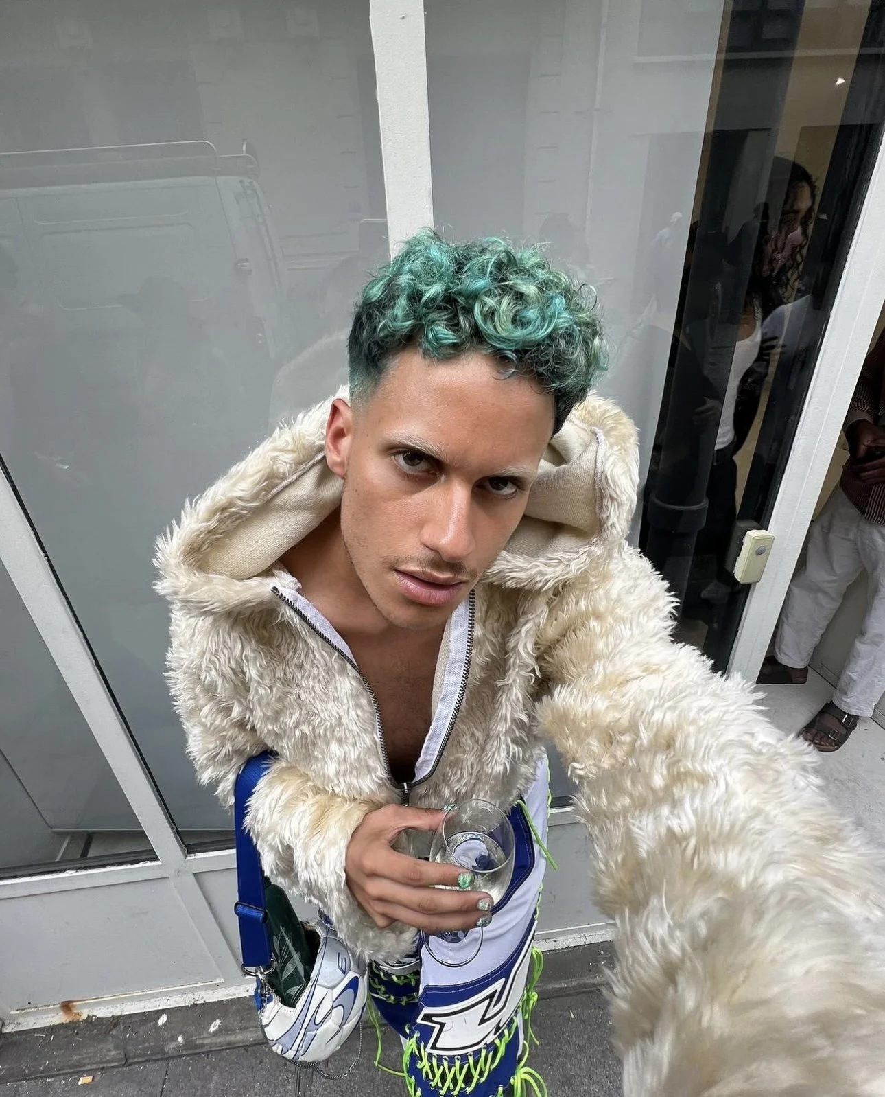 Un homme avec des cheveux bouclés de couleur turquoise, portant une veste en fausse fourrure crème, tenant un verre à la main, photographié à partir d'une prise de vue en hauteur.