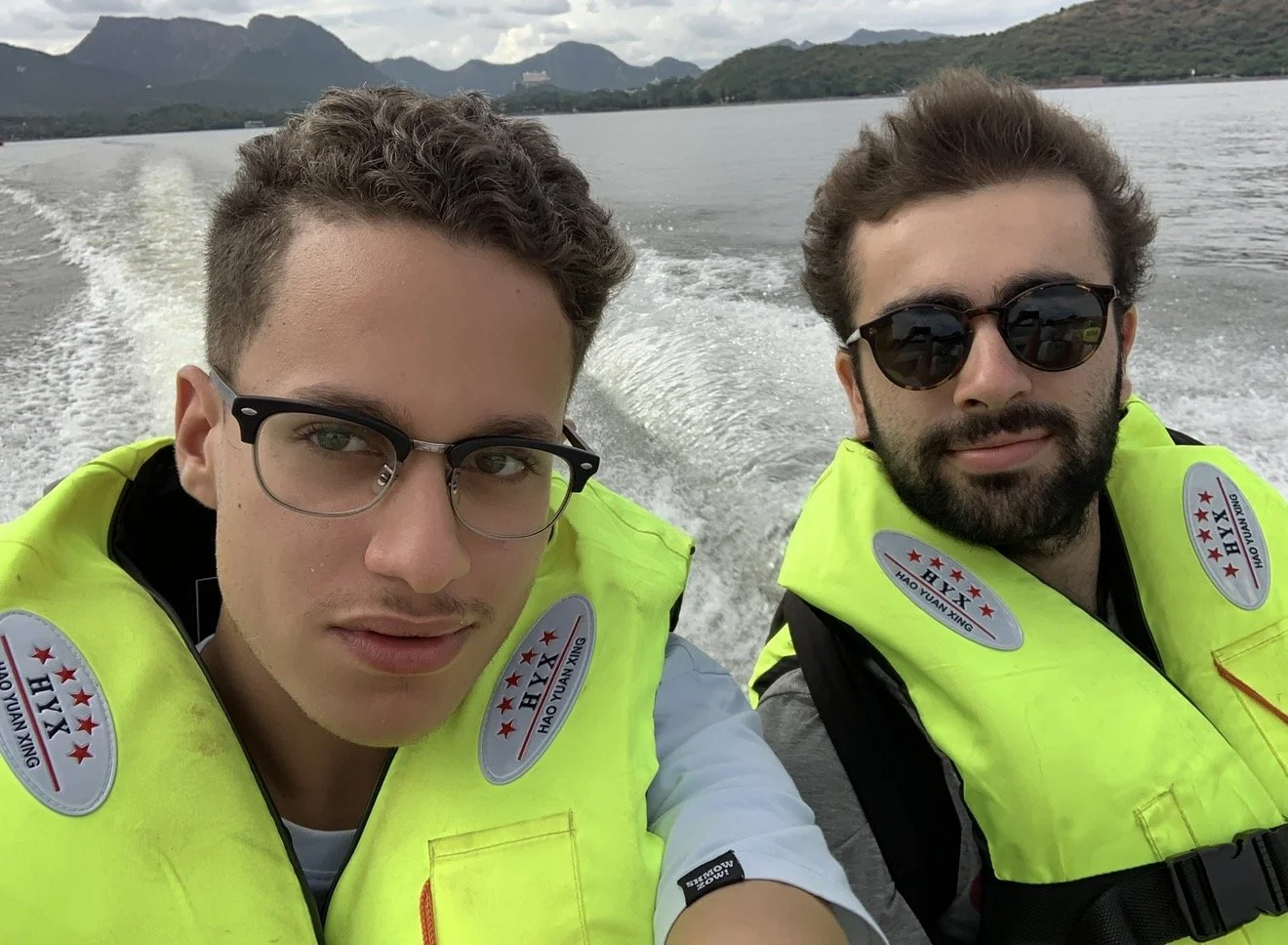Deux hommes portant des gilets de sauvetage jaunes prennent un selfie sur un bateau, avec des montagnes en arrière-plan.