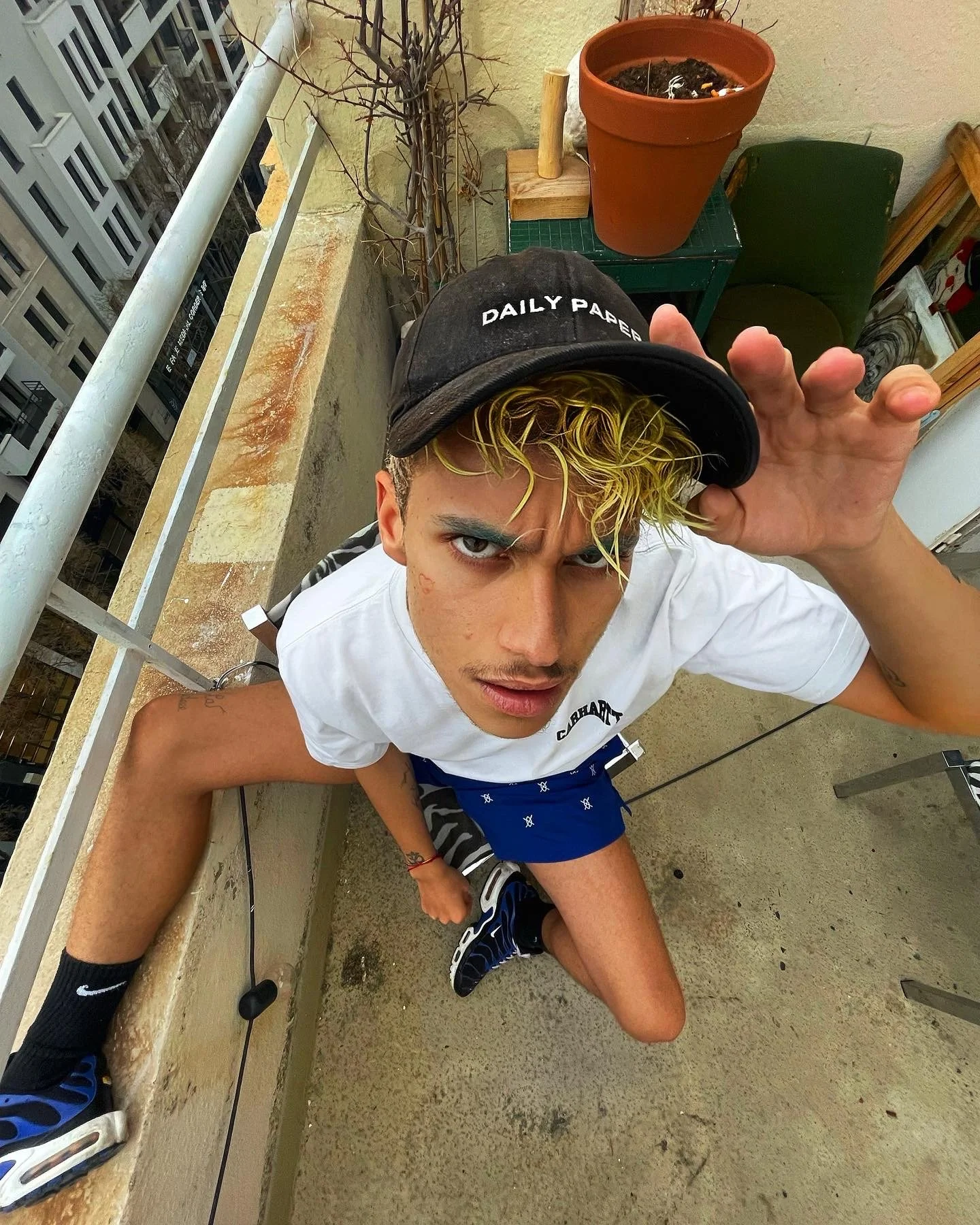 Jeune homme avec cheveux blonds et bouclés portant une casquette noire écoutant un appel, assis sur un balcon avec des plantes et des meubles en arrière-plan.