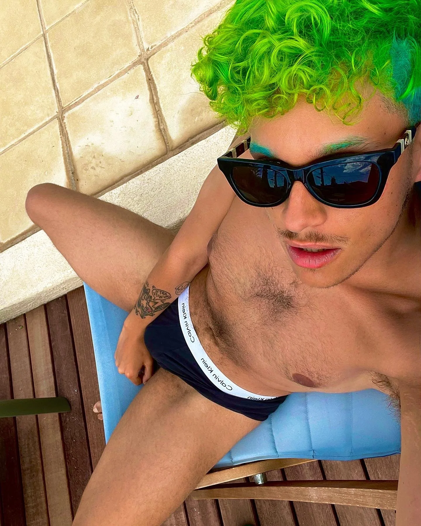 Un jeune homme ombragé porte des lunettes de soleil noires, a des cheveux bouclés verts fluorescents, est allongé sur un drap bleu en position semi-allongée sur un sol en bois avec un mur beige en arrière-plan.