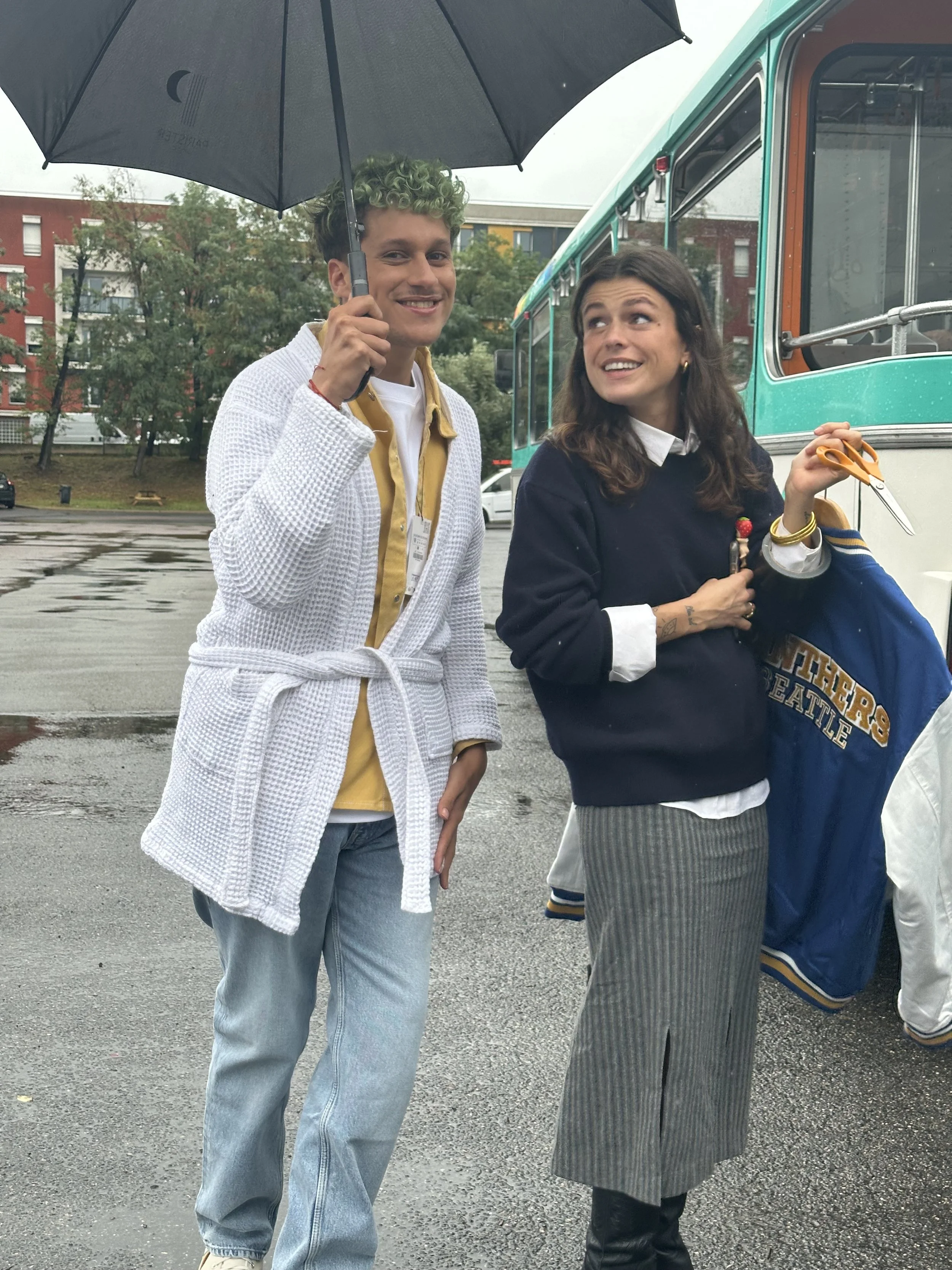 Deux jeunes femmes souriantes sous un parapluie, une d'elles tenant des ciseaux, debout devant un bus à l'extérieur un jour pluvieux.