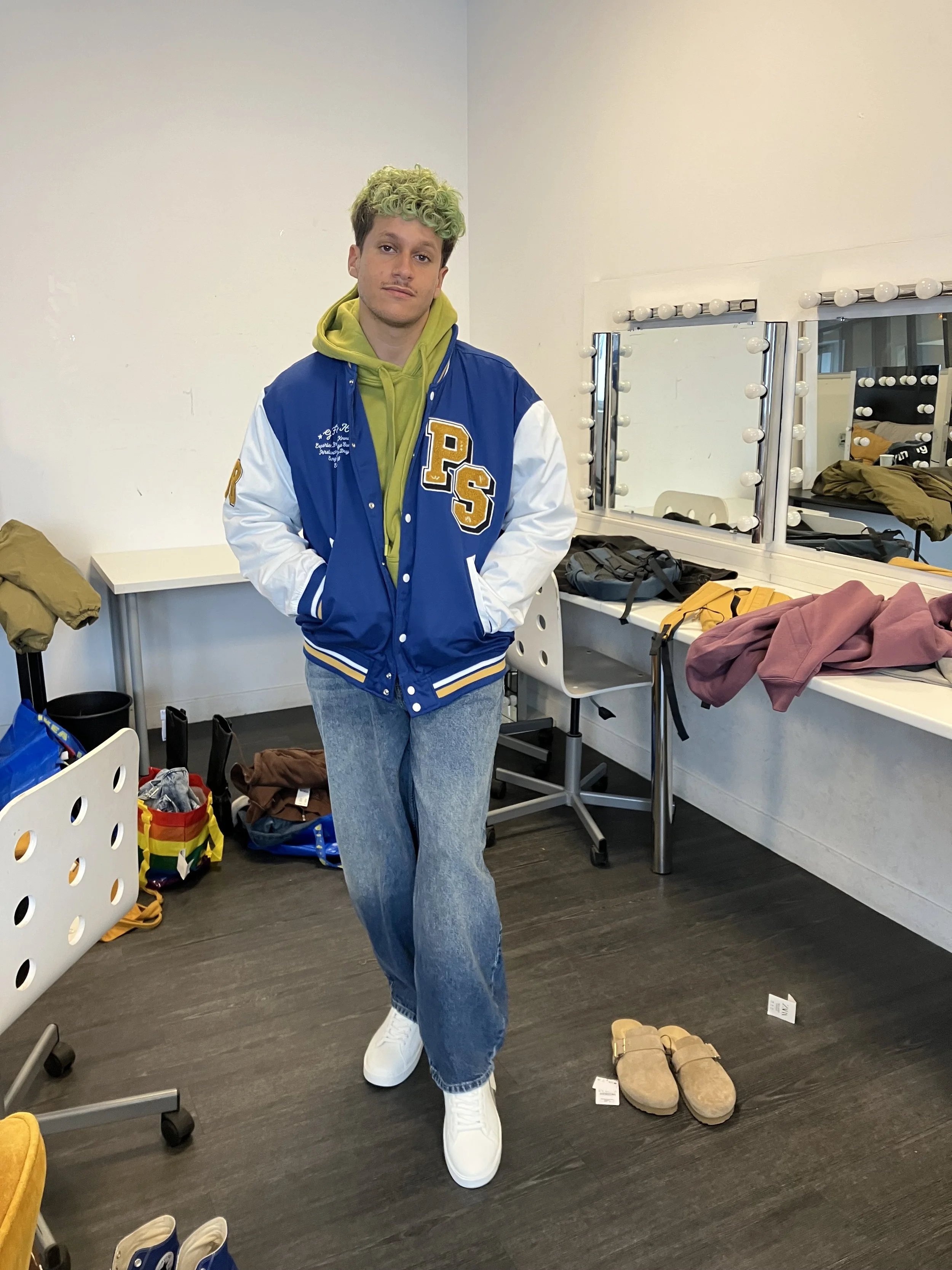 Jeune homme avec des cheveux bouclés verts portant un blouson de sport bleu et blanc, un sweat à capuche vert, et un jean bleu, dans une salle de vestiaire avec des miroirs et des vêtements autour.