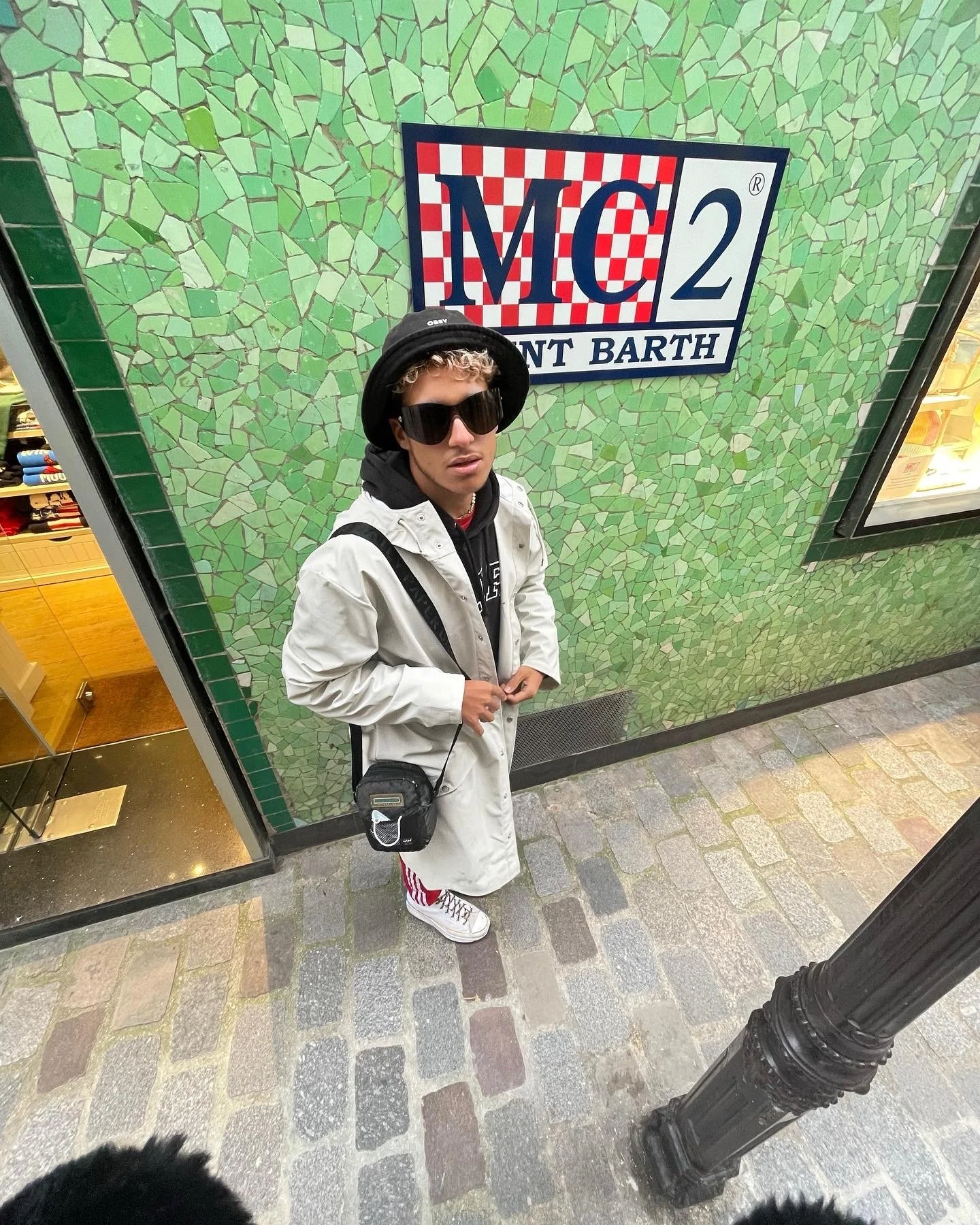 Une personne portant des lunettes de soleil, un chapeau noir, un manteau blanc, et un sac à bandoulière, se tenant devant un mur avec un panneau qui indique 'MC2 Saint Barth' dans un environnement urbain.
