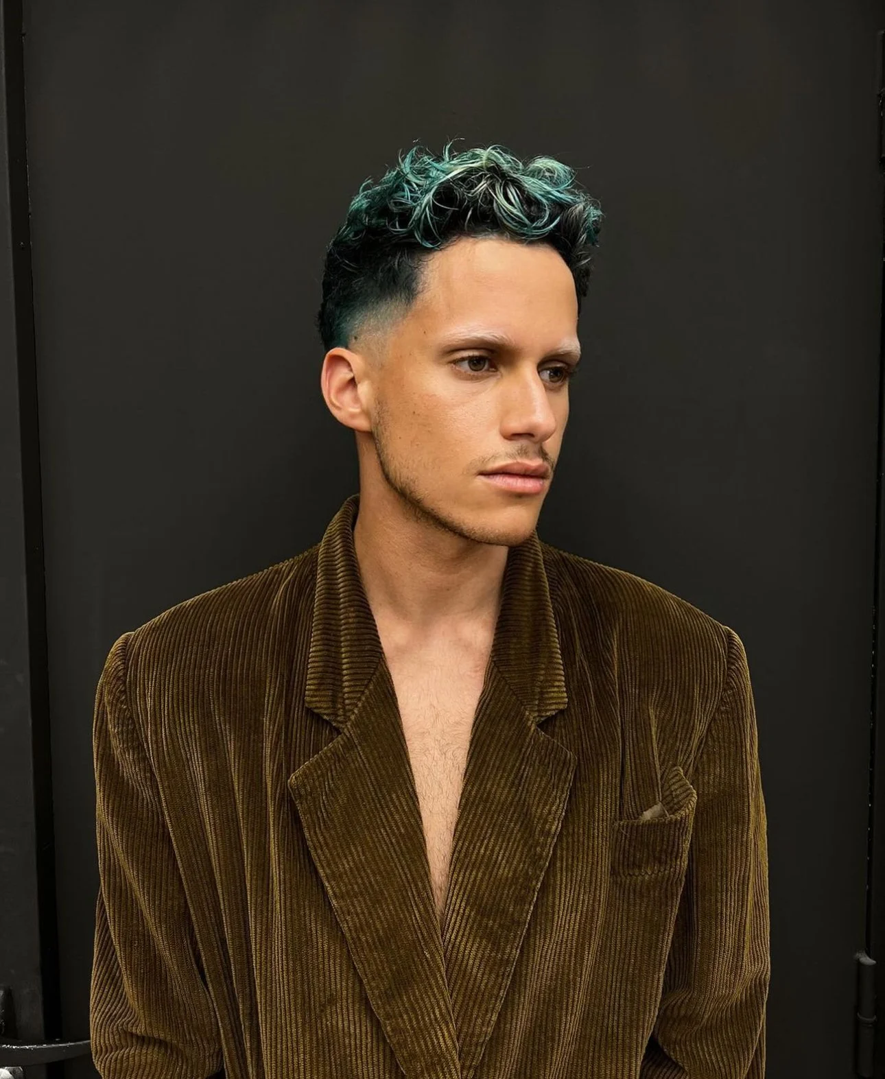 Un jeune homme avec des cheveux coiffés en boucles colorées en turquoise et noir, portant un costume en velours marron, posé devant un fond noir.