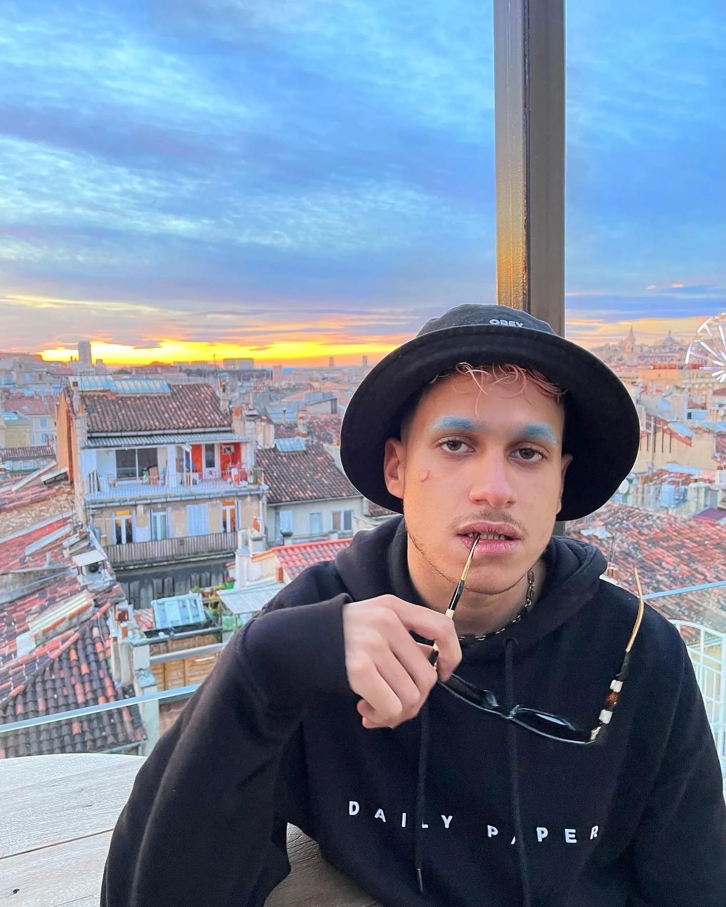 Un jeune homme portant un chapeau noir et un sweat noir, avec du maquillage bleu sur les yeux, tenant un pinceau près de ses lèvres, assis devant une vue sur la ville au coucher du soleil, avec un ciel coloré et des toits en tuiles.