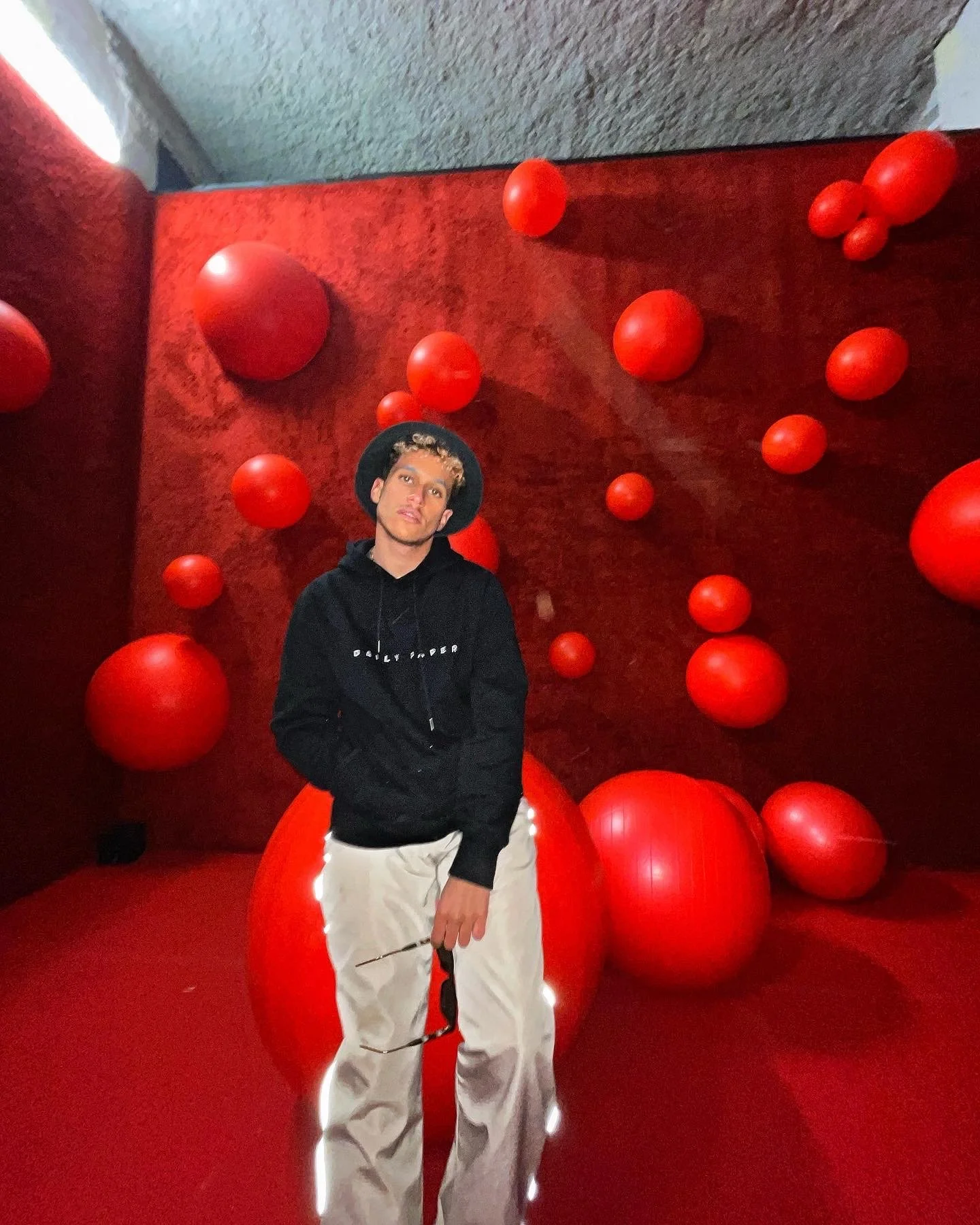 Jeune homme avec un chapeau noir, chemise noire, et pantalon beige, posant dans un décor avec de grands ballons rouges suspendus contre un mur rouge.