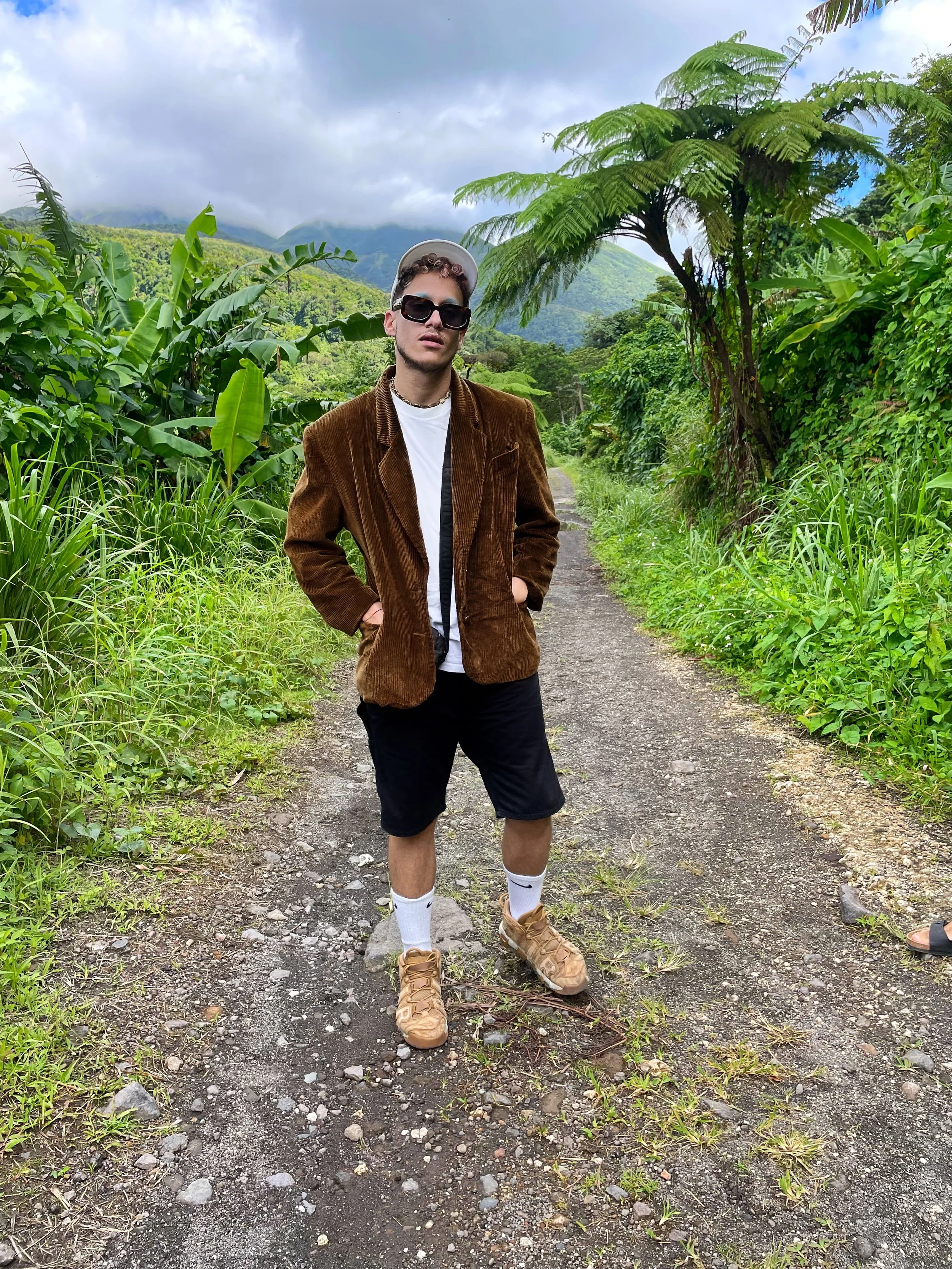 Jeune homme avec un blazer marron, short noir, baskets beige, chaussettes blanches, portant des lunettes de soleil, dans une jungle ou forêt dense, sur un chemin, avec des montagnes et des nuages à l'horizon.