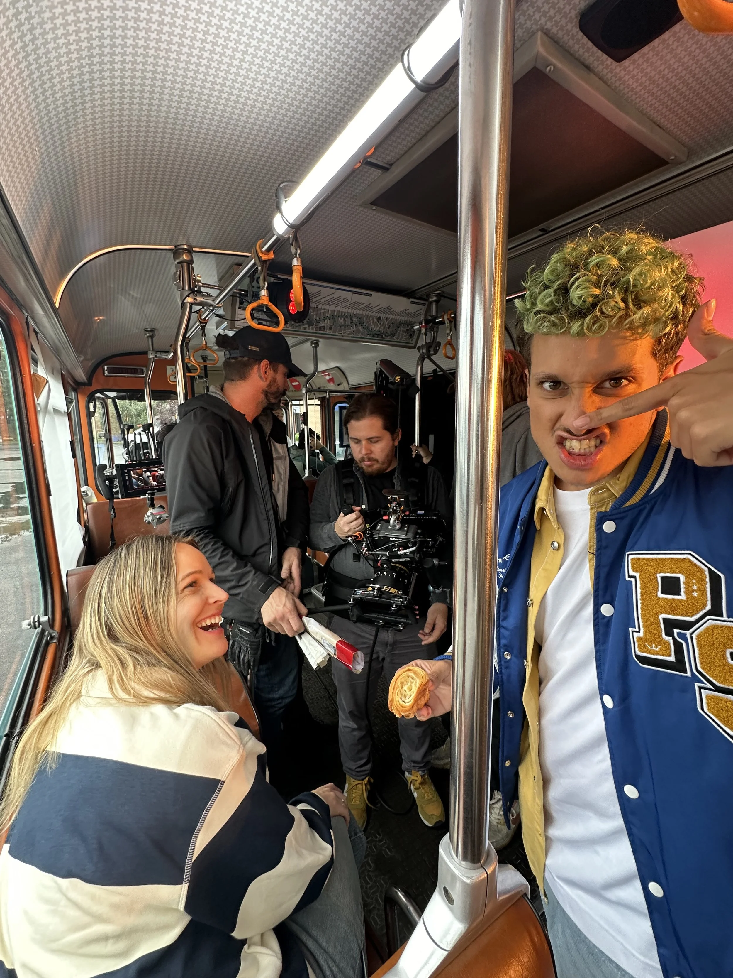 Deux jeunes personnes dans un bus, une femme souriante assise, un homme avec des cheveux bouclés pointant vers lui, entourés de l'équipe de tournage.