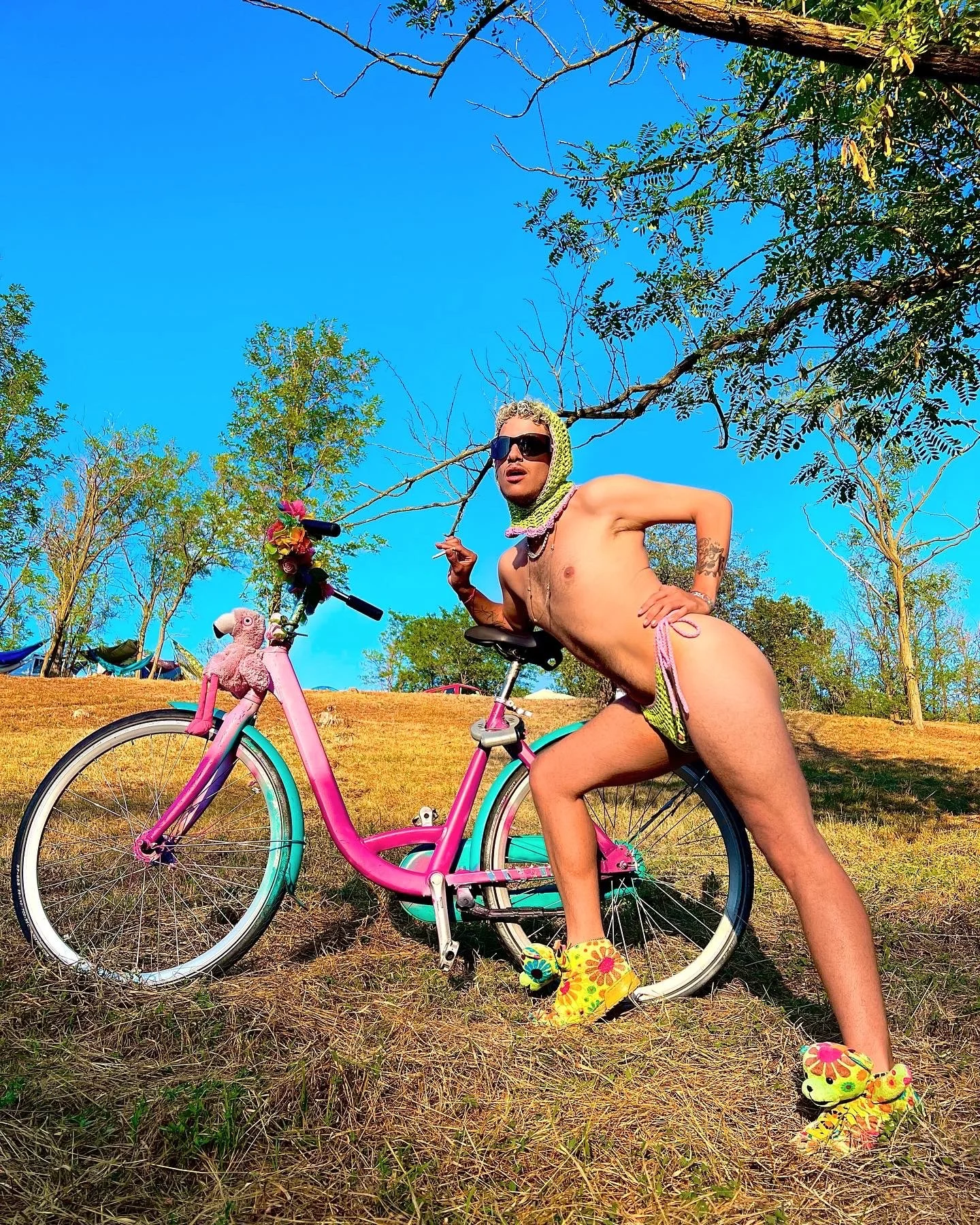 Une personne nue porte des chaussures colorées, des lunettes de soleil, un foulard sur la tête, pose à côté d'un vélo rose décoré avec un ballon en peluche de flamant rose, dans un paysage extérieur avec des arbres et un ciel bleu.