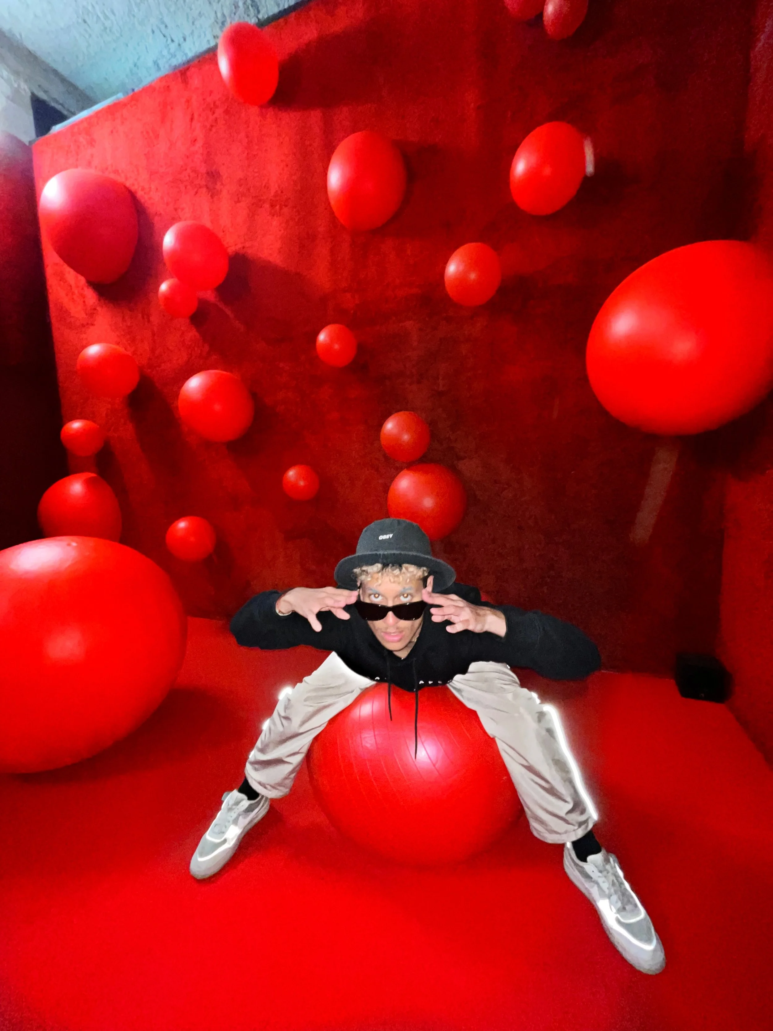 Une personne posant avec des ballons rouges dans une pièce rouge, portant des lunettes de soleil, un chapeau gris et des vêtements décontractés.