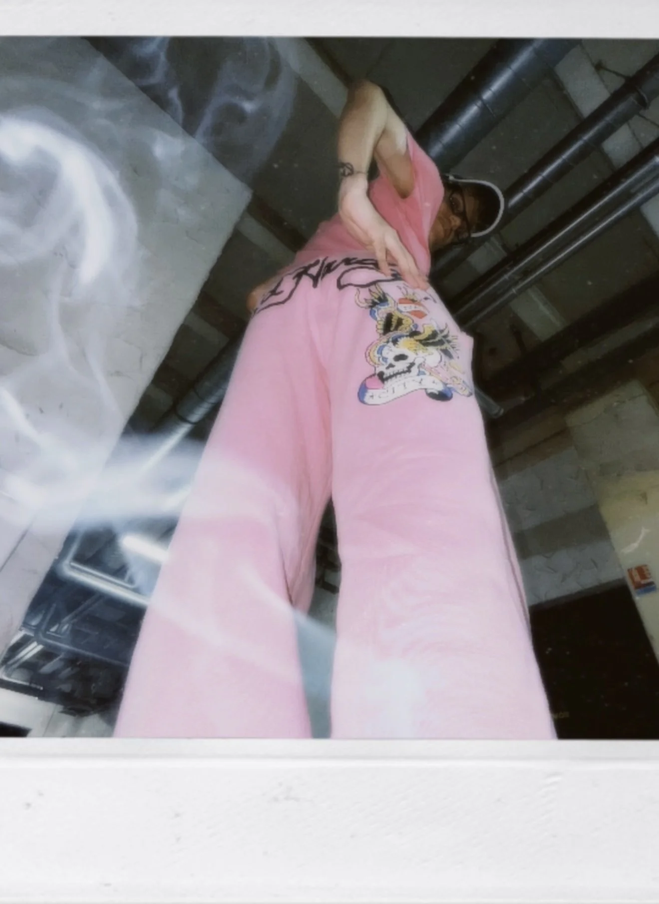 Une personne portant un pantalon rose avec un design de crâne et un t-shirt rose, vue du dessous, dans un environnement industriel.
