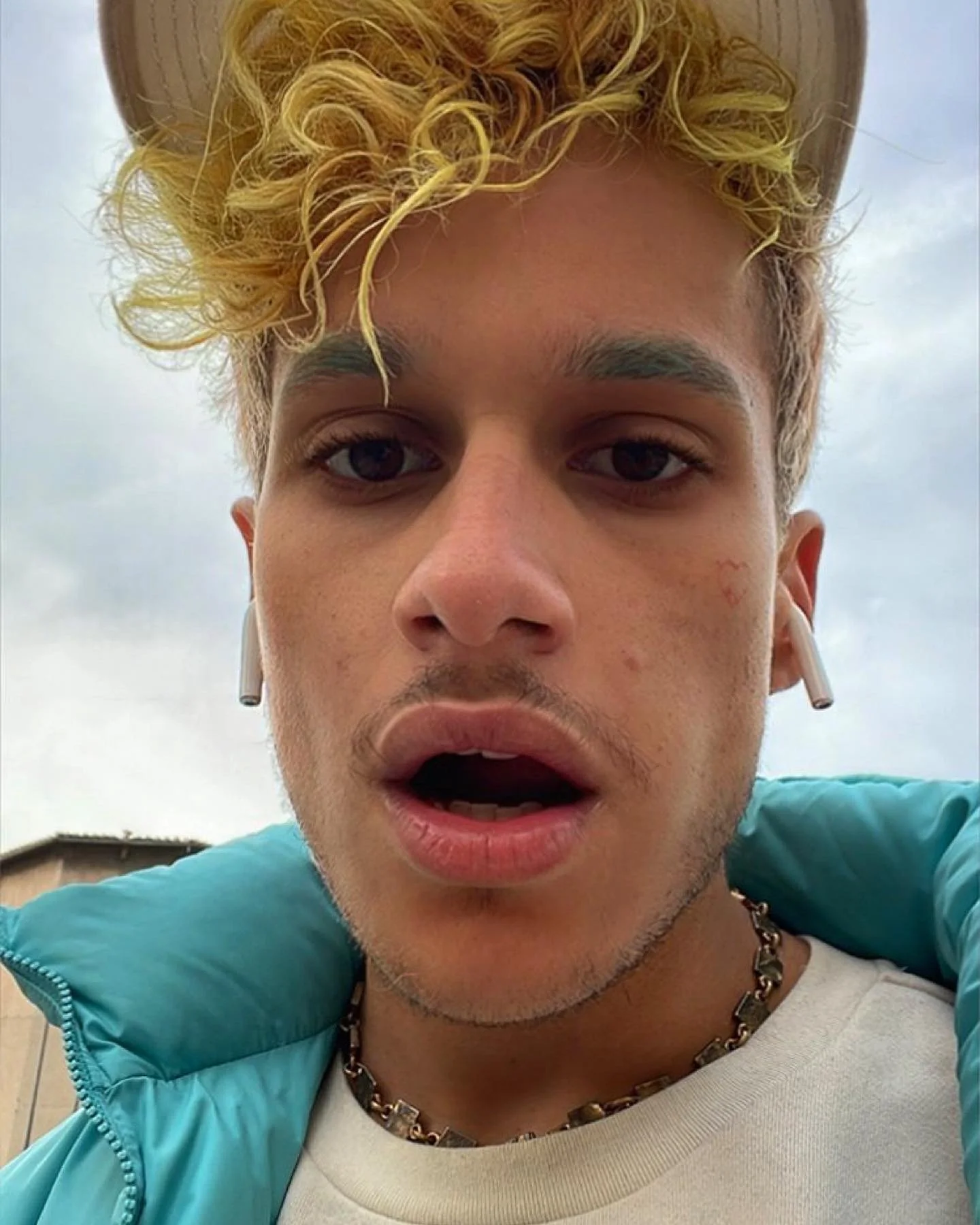 Jeune homme aux cheveux blonds bouclés portant des écouteurs sans fil, un collier en chaîne, un sweat bleu et un t-shirt blanc, faisant une expression de surprise ou de curiosité à l'extérieur par temps nuageux.