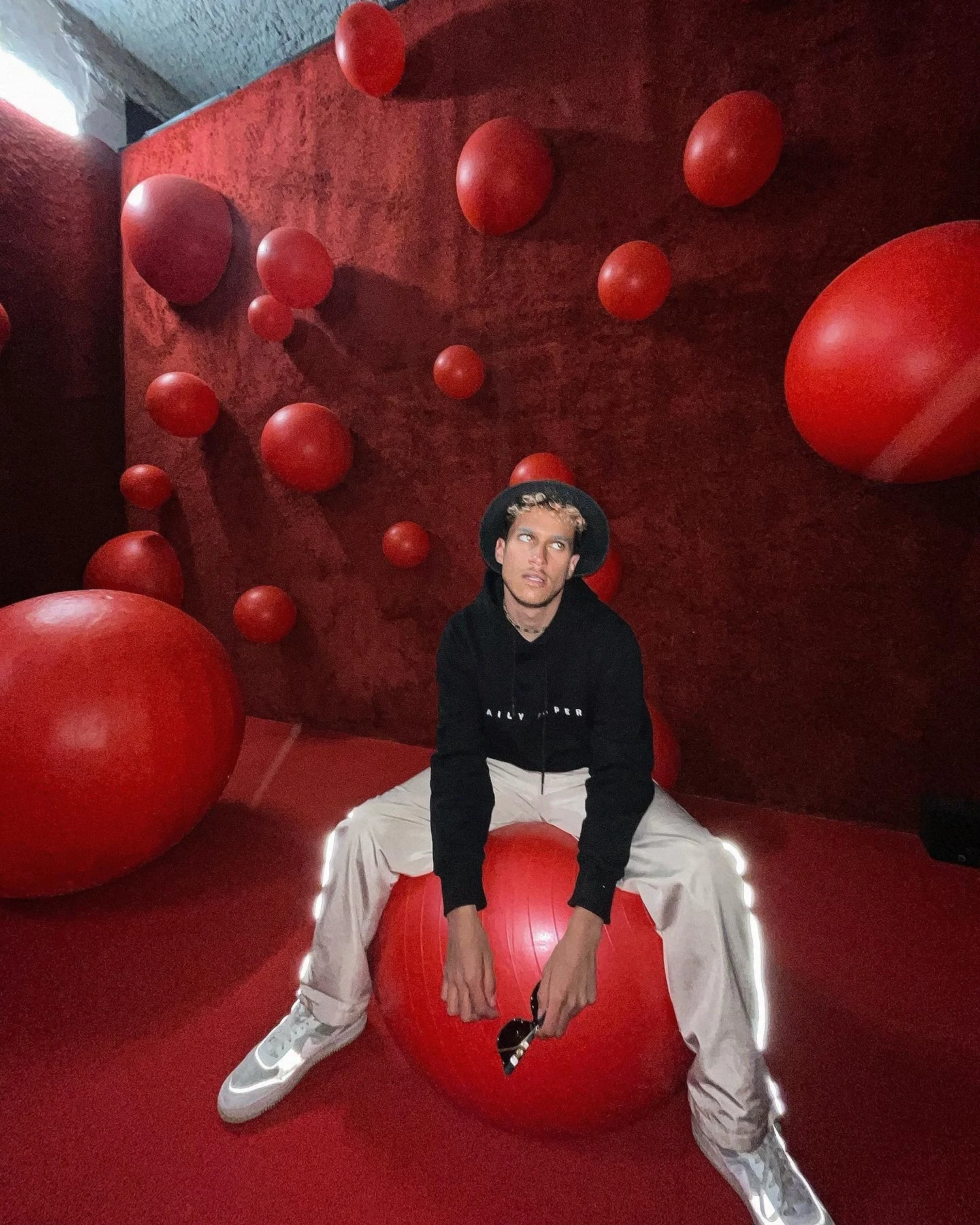 Une jeune personne assise sur un ballon rouge dans une pièce décorée avec de nombreux autres ballons rouges, le mur derrière elle est rouge foncé.