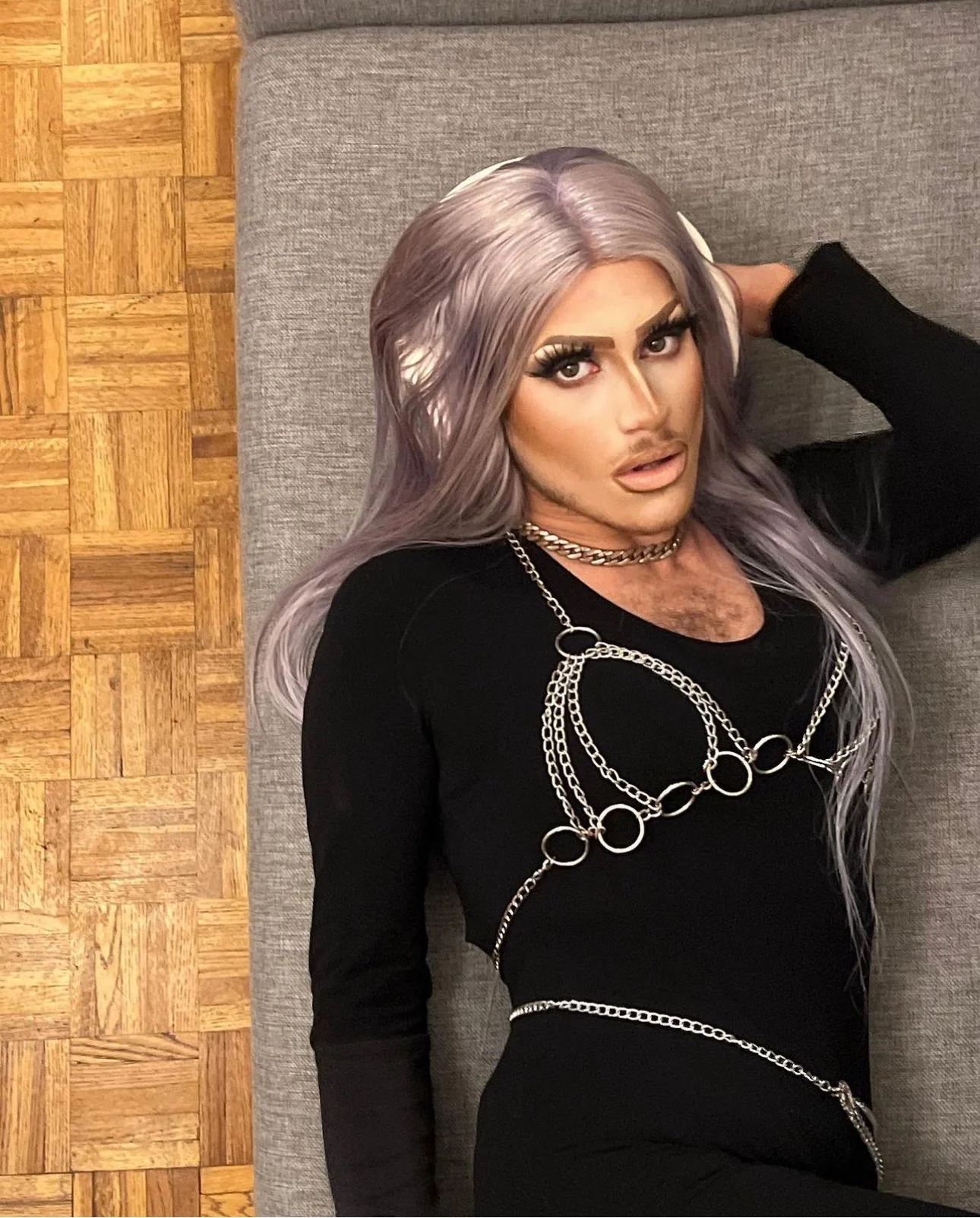Une personne aux cheveux longs et violets, portant un maquillage élaboré, un chemisier noir et des chaînes en or, repose sur un canapé en tissu gris avec un plancher en bois en arrière-plan.